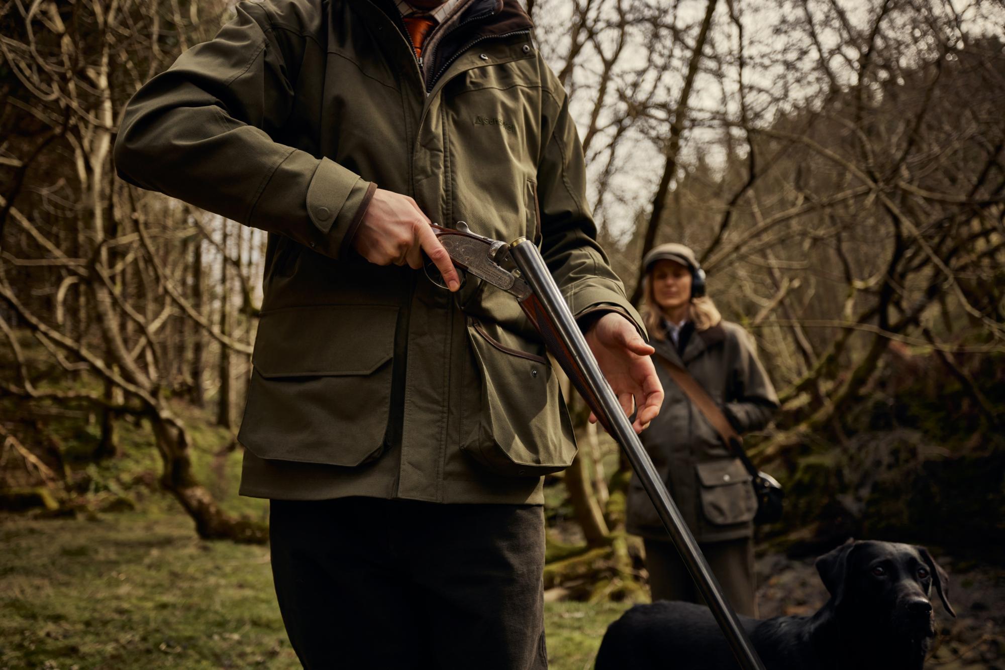 Two people in outdoor clothing with a rifle in a forest setting