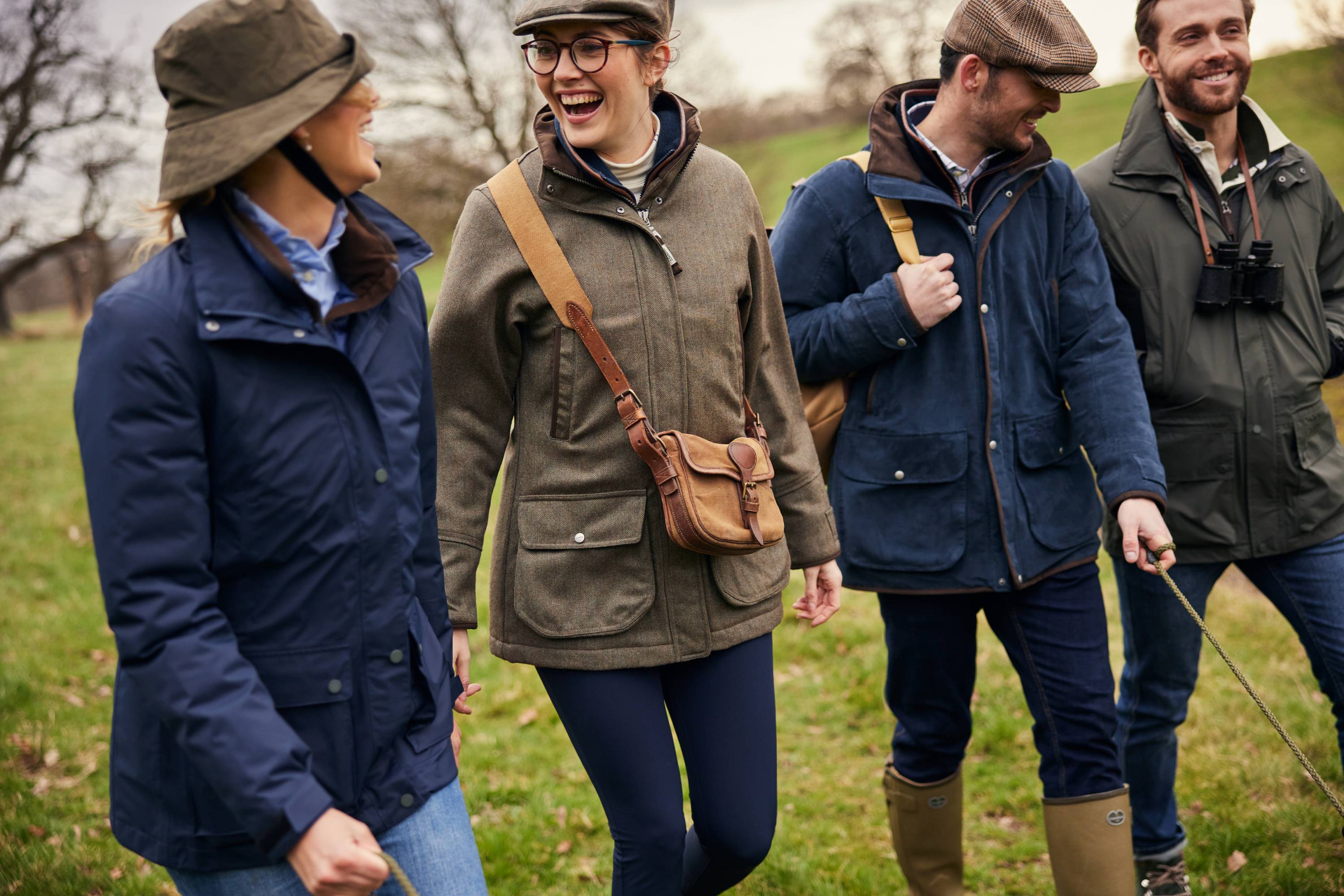 Four friends in country-style outdoor clothing walk through a grassy countryside, laughing and chatting. They wear tweed jackets, waxed coats, and boots, with one holding a leash and another carrying binoculars.