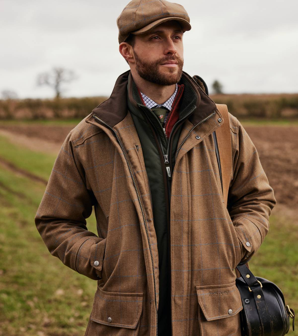 A man wearing a tweed jacket, green gilet, checked shirt, tie and tweed cap in a field. He is standing with both hands in his pockets and he is looking away from the camera.