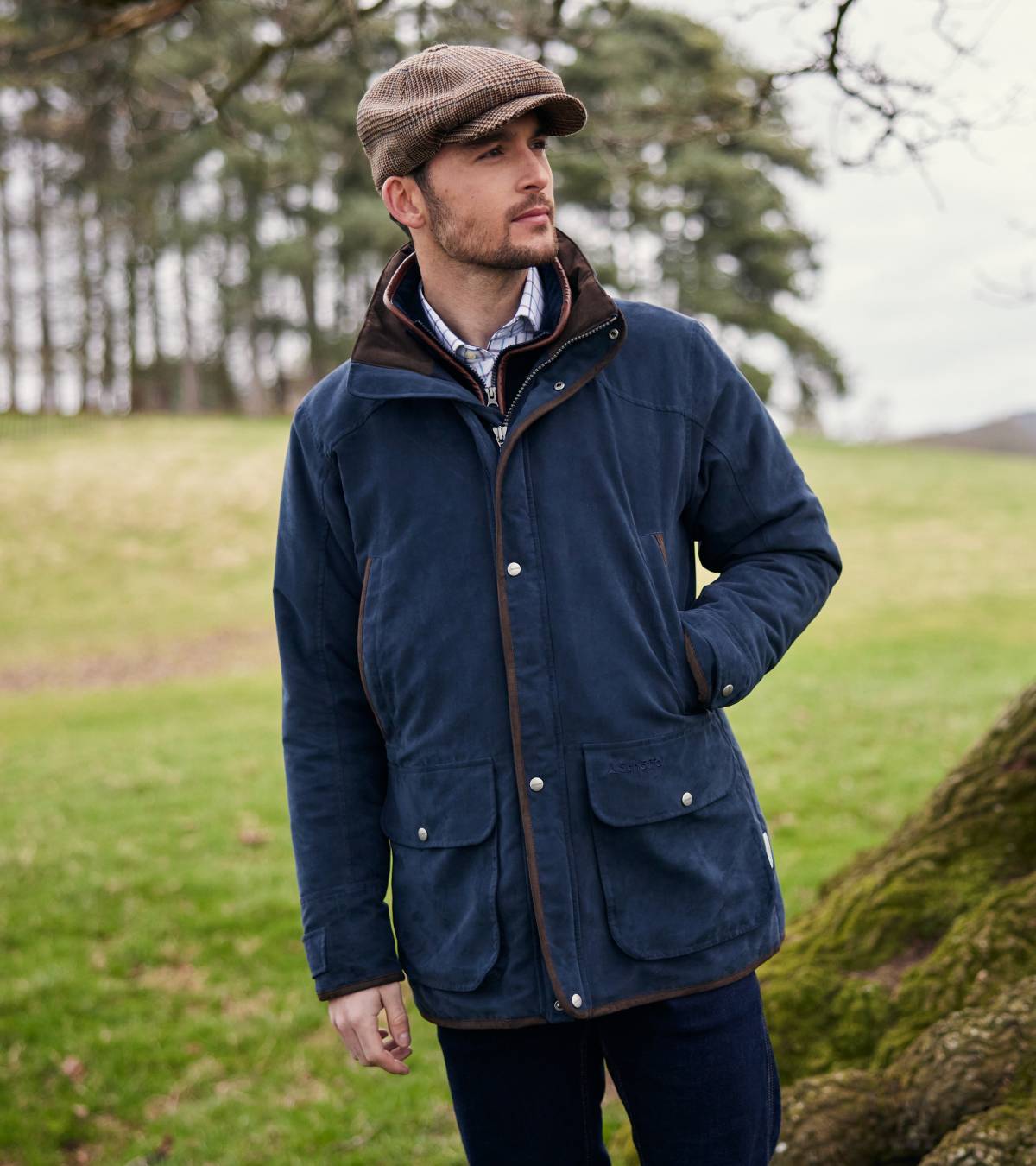 A man wearing a navy jacket, checked shirt and tweed cap in a field. He is standing with one hand in his pocket and he is looking away from the camera.