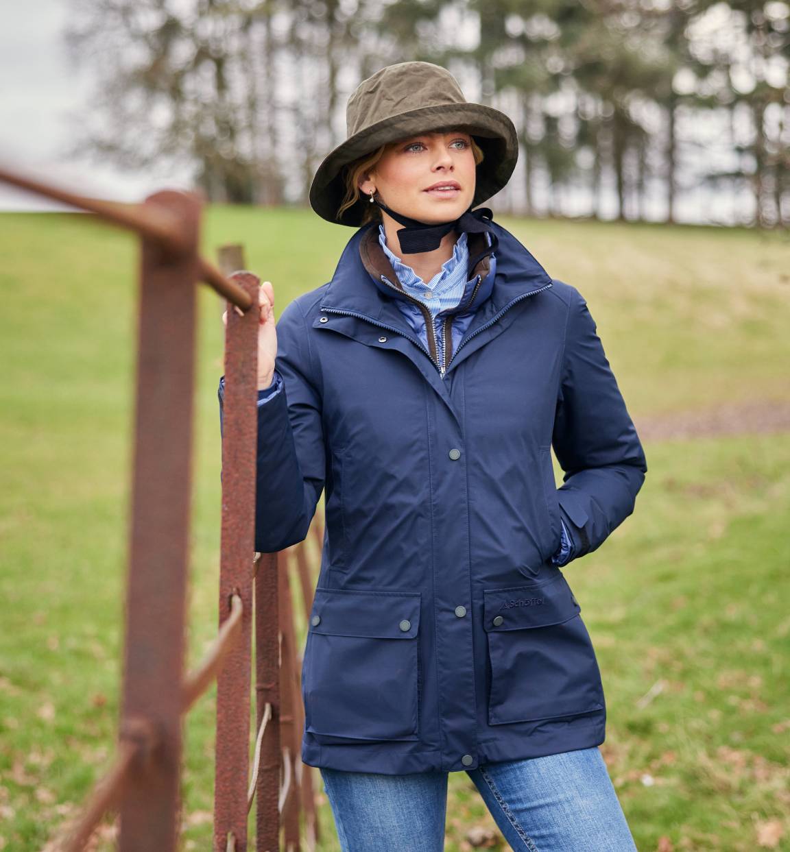 A lady wearing a navy jacket, checked shirt and tweed cap in a field, leaning against a fence. She is standing with one hand in her pocket and he is looking away from the camera.