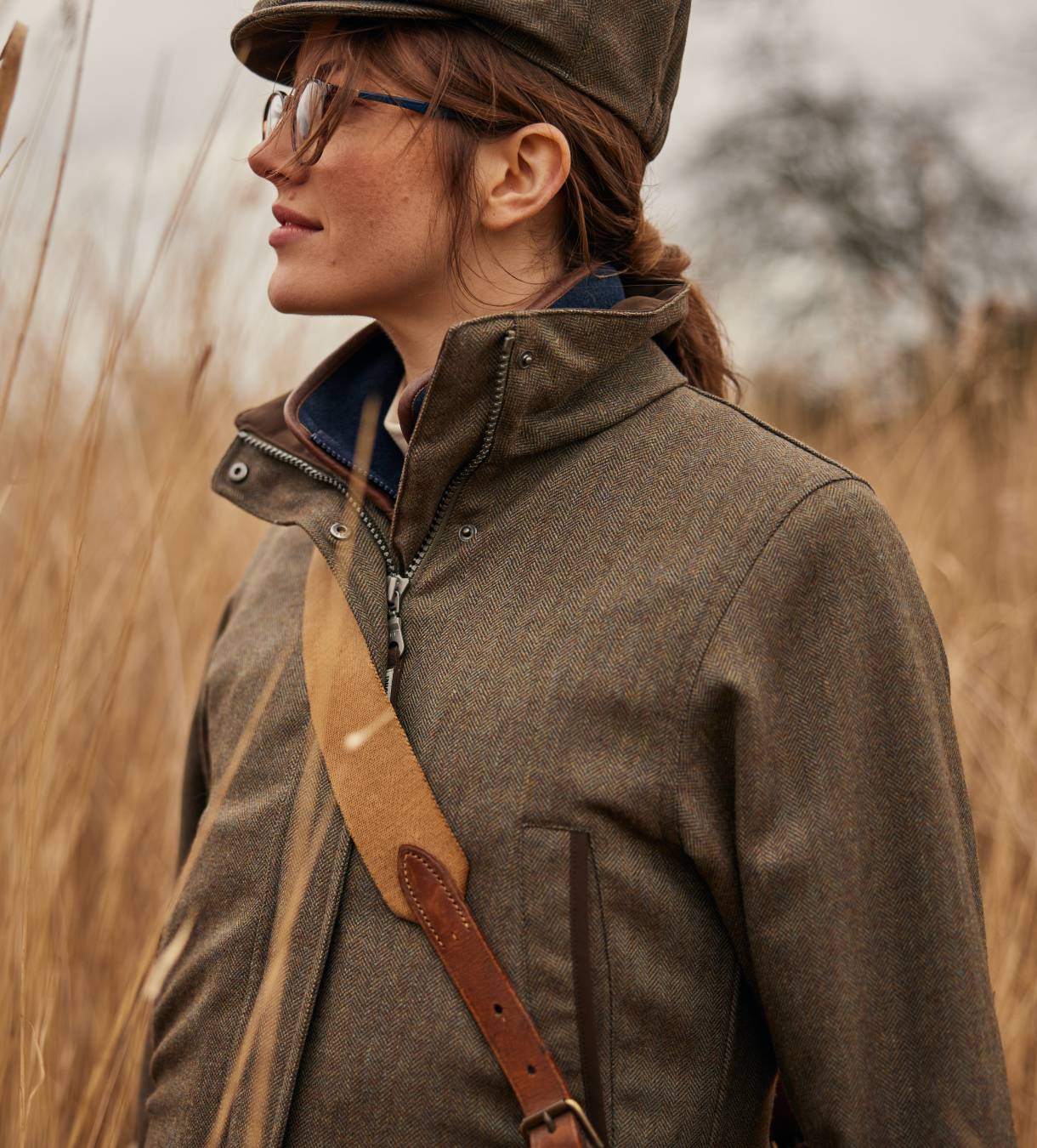 A woman wearing a tweed jacket, navy gilet and tweed cap in a field. She is smiling looking away from the camera.