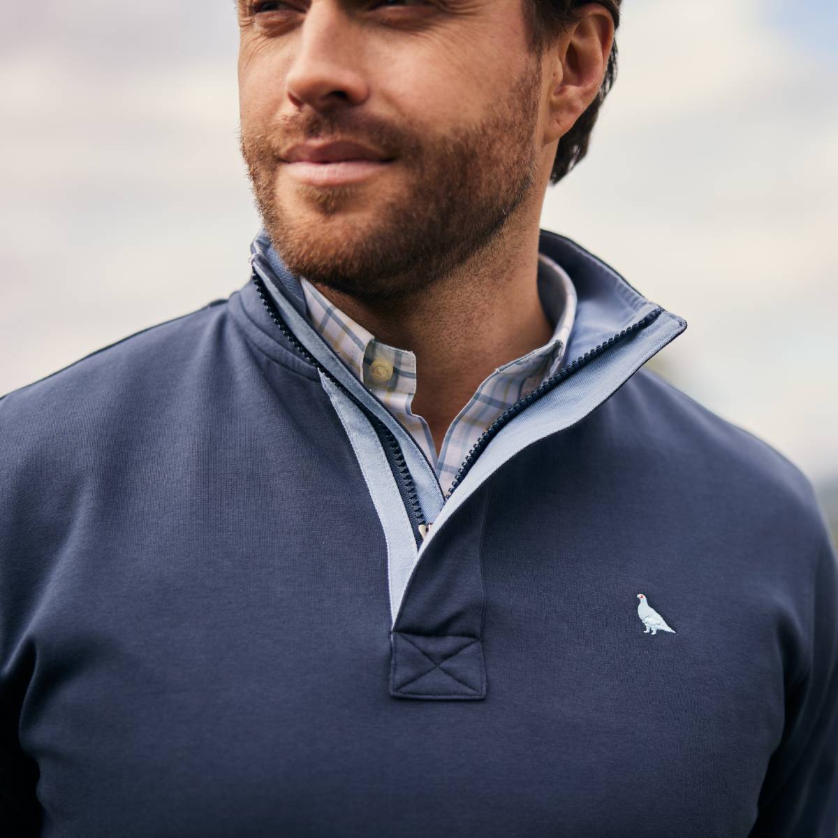 Close up image of a blue sweatshirt with blue and pink checked shirt underneath. Image highlights the contrasting collar and light blue embroidered bird logo on the chest.