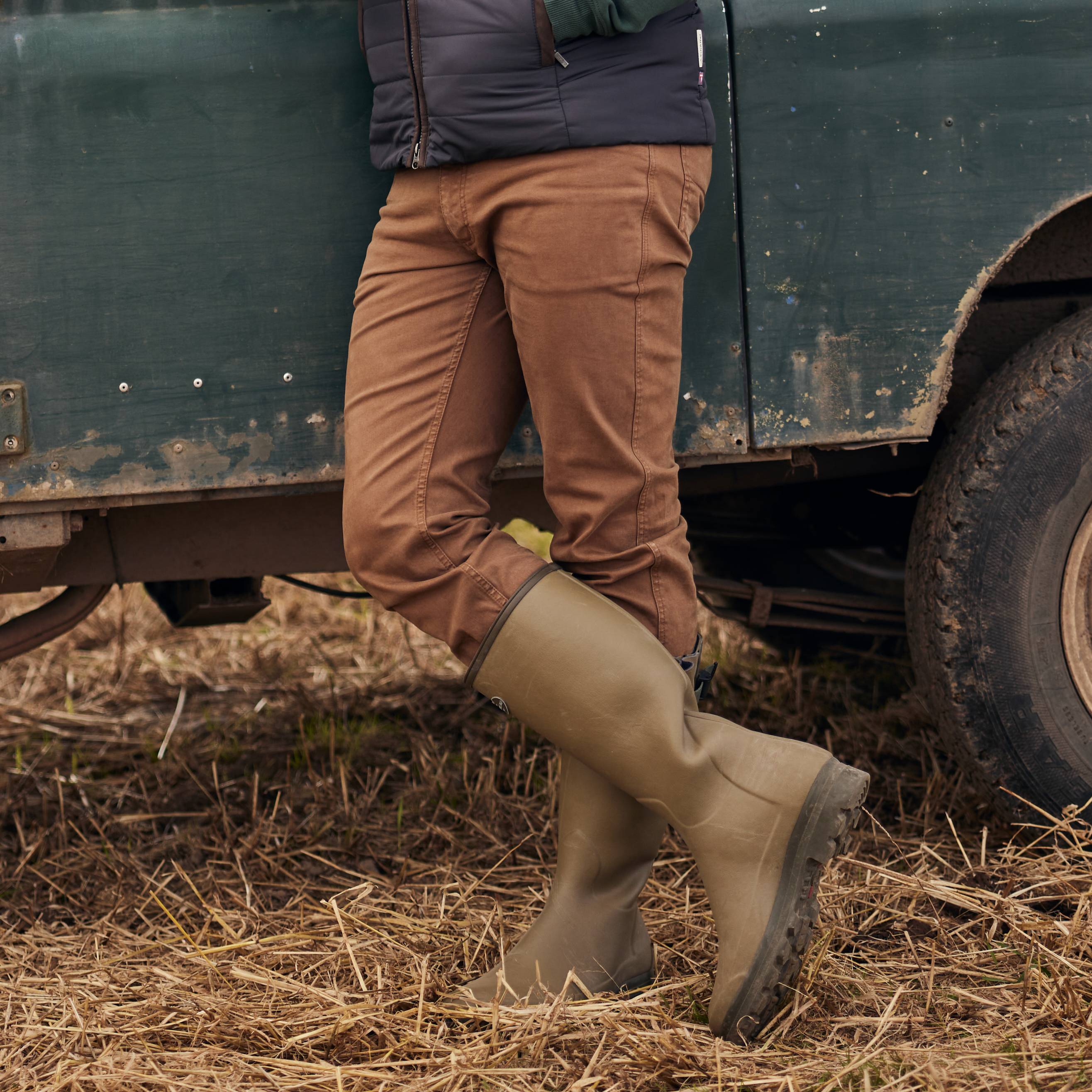 A close-up of a person's lower body leaning against a vintage green Land Rover. They are wearing brown trousers, a navy blue jacket, and tall olive-green rubber Wellington boots with a rugged sole. The ground is covered in dry grass and mud, suggesting an outdoor, rural setting.