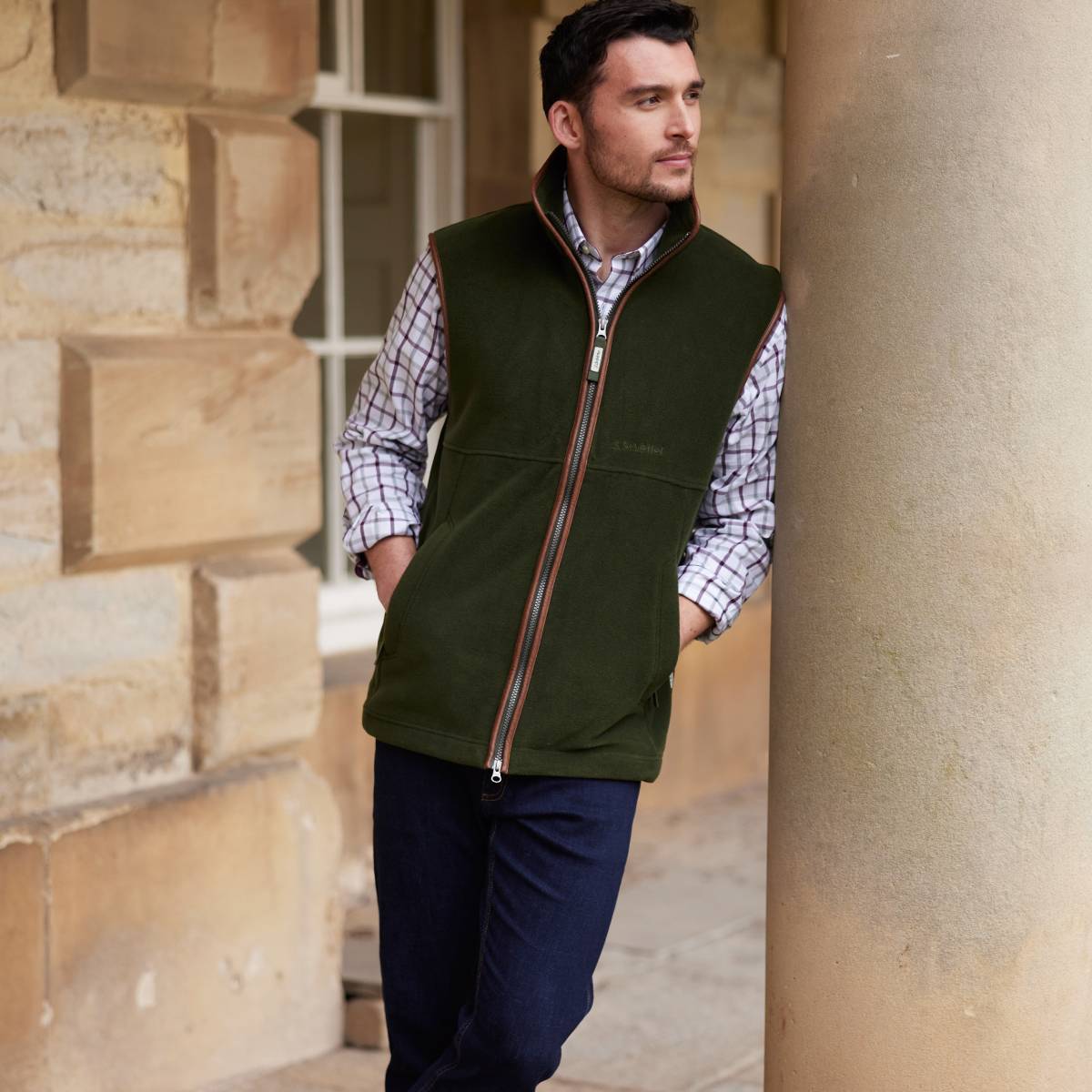 A man wearing a green gilet over a checked shirt and dark blue jeans. He is standing in a relaxed pose looking away from the camera.