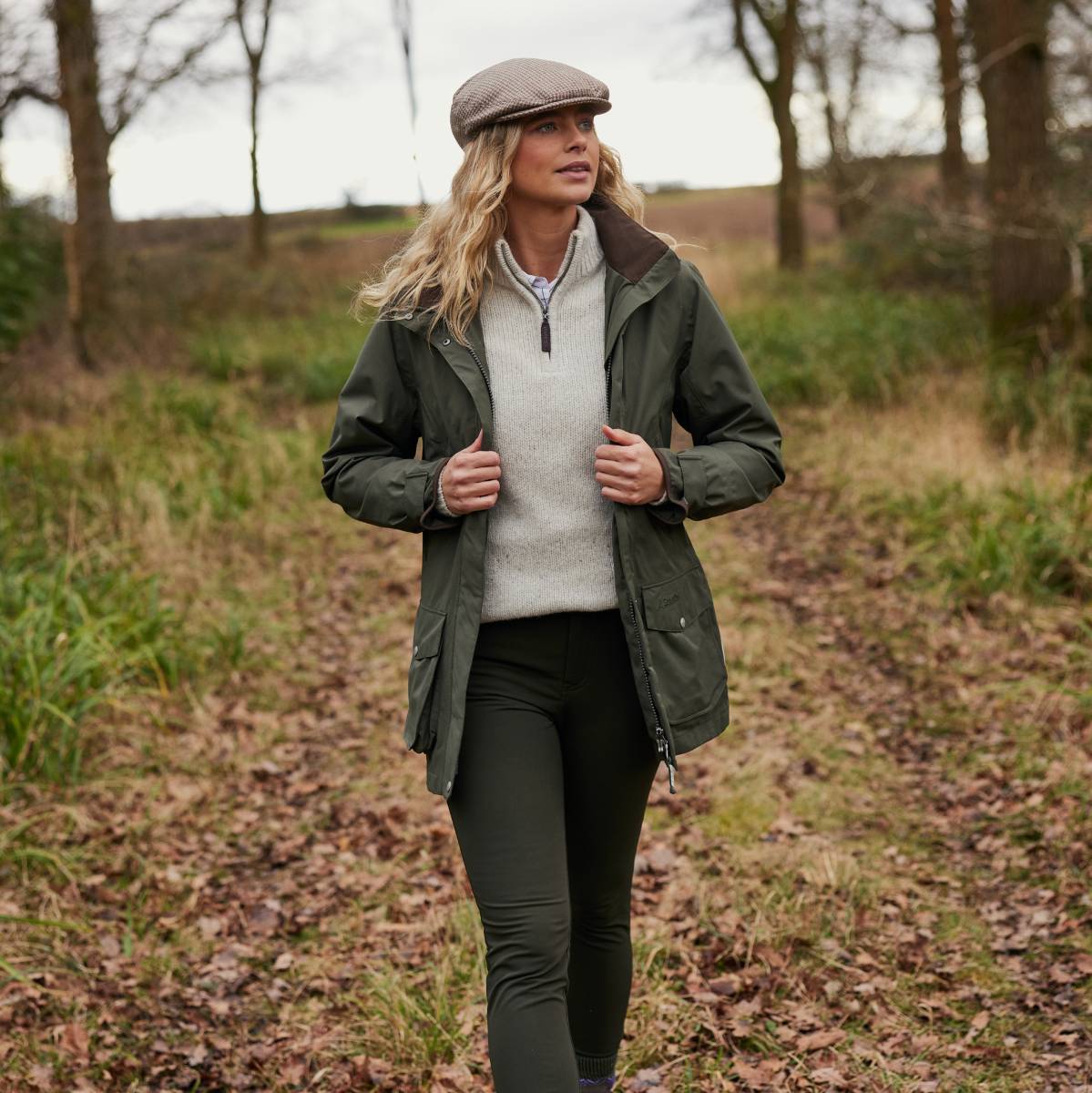 An image of a lady walking in woodland wearing a green jacket, tweed cap and beige quarter zip jumper.