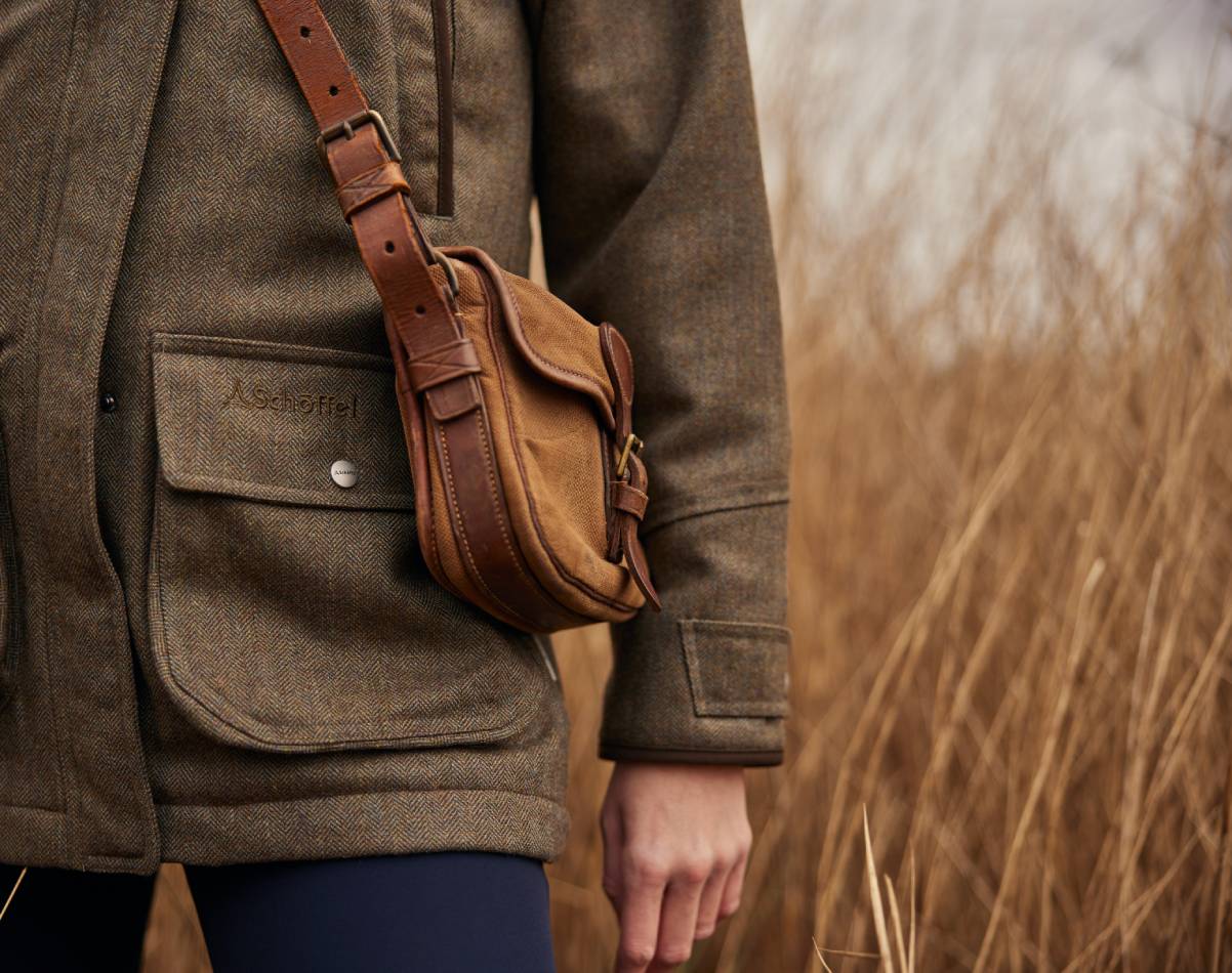 Close up image of a tweed jacket with a leather cross body bag. Image highlights the pockets and adjustable cuffs.