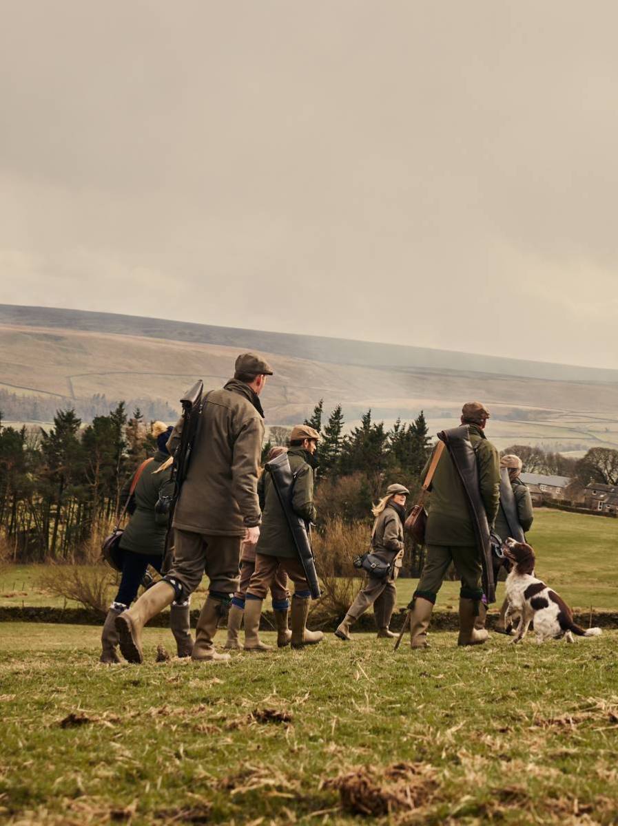 A group of people and dogs walking in a field wearing countrywear.