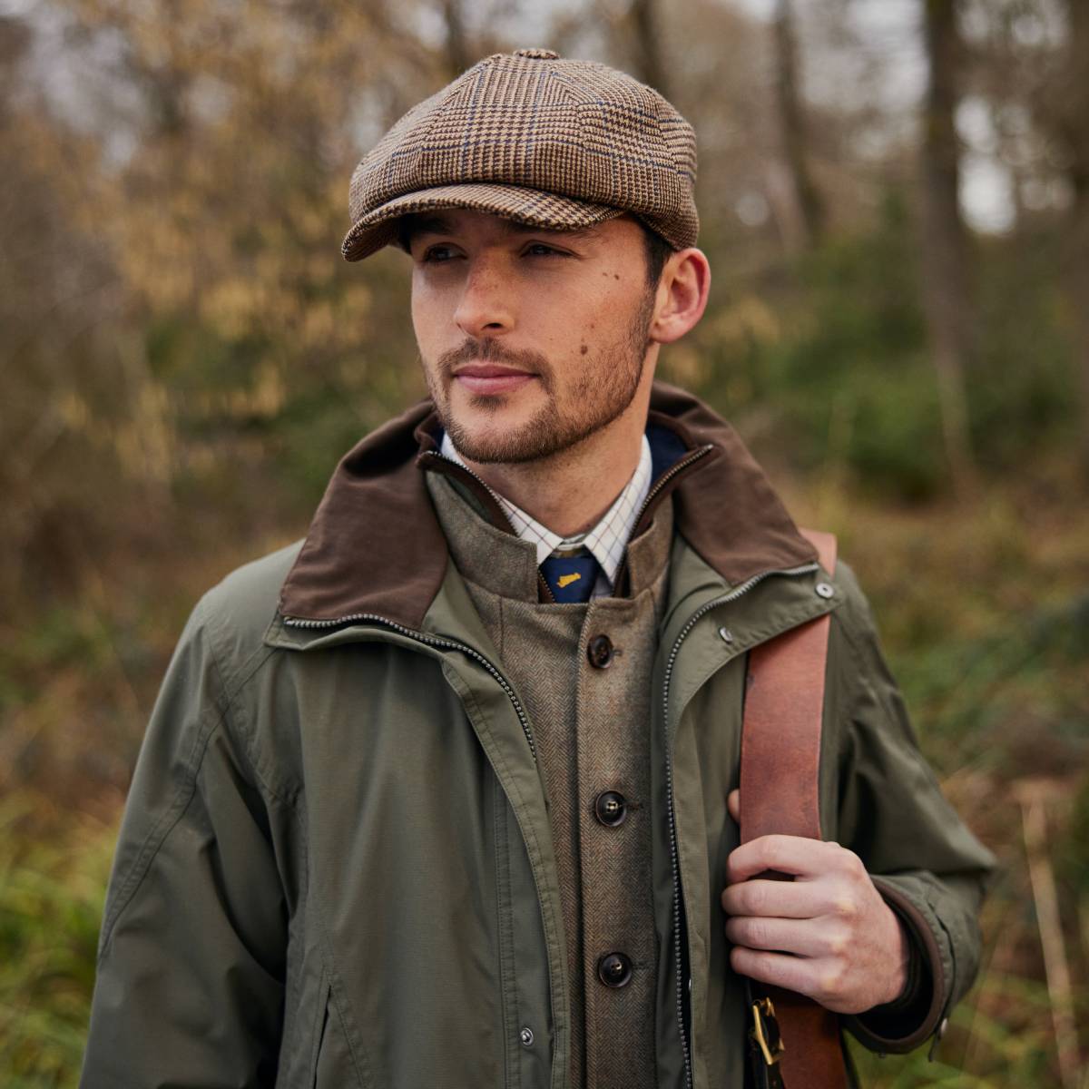An image of a man wearing a tweed waistcoat over a checked shirt and blue tie. He is wearing a green country jacket and tweed cap. He is smiling looking away from the camera.