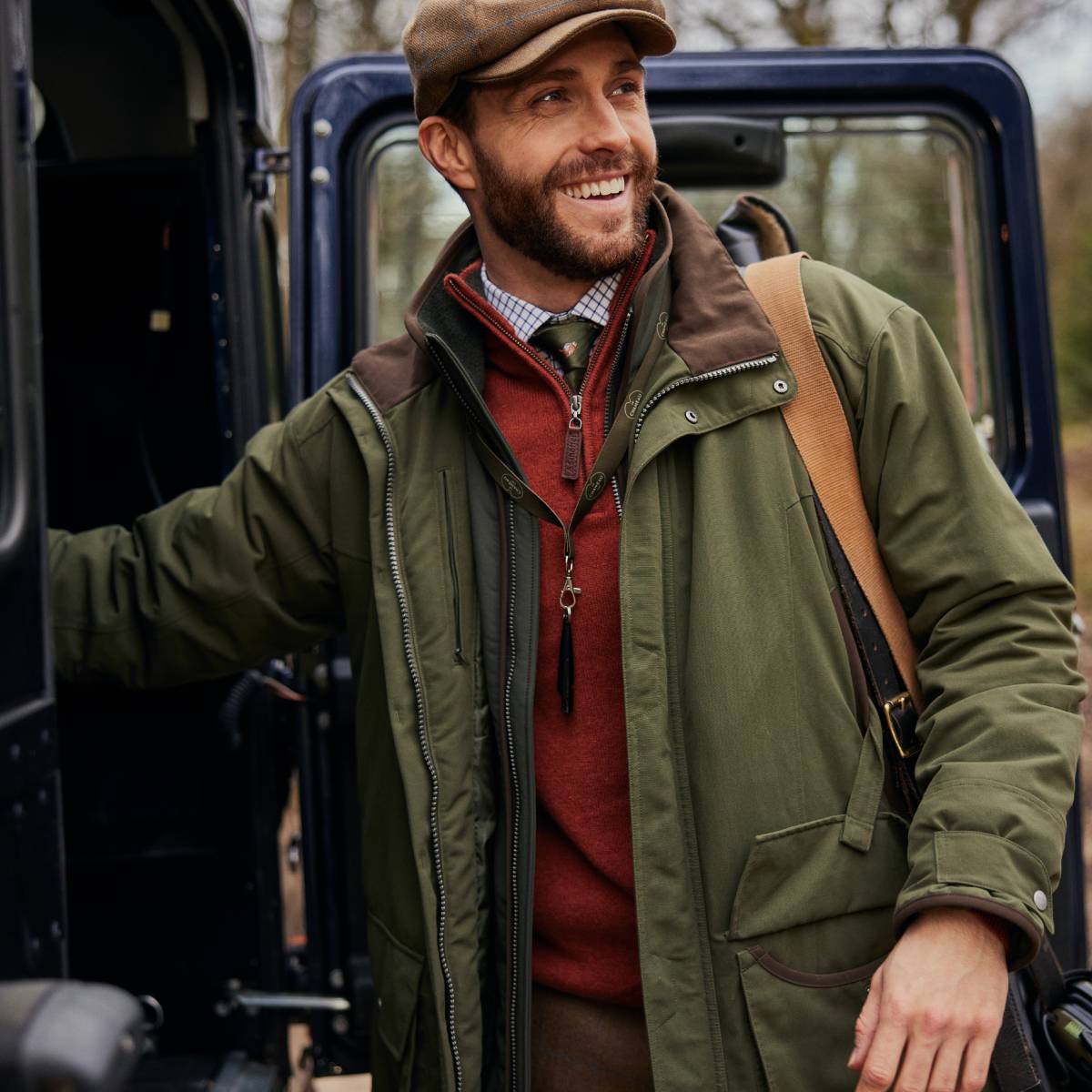An image of a man wearing a red quarter zip jumper over a checked shirt and green tie. He is wearing a green country jacket and tweed cap. He is smiling looking away from the camera.