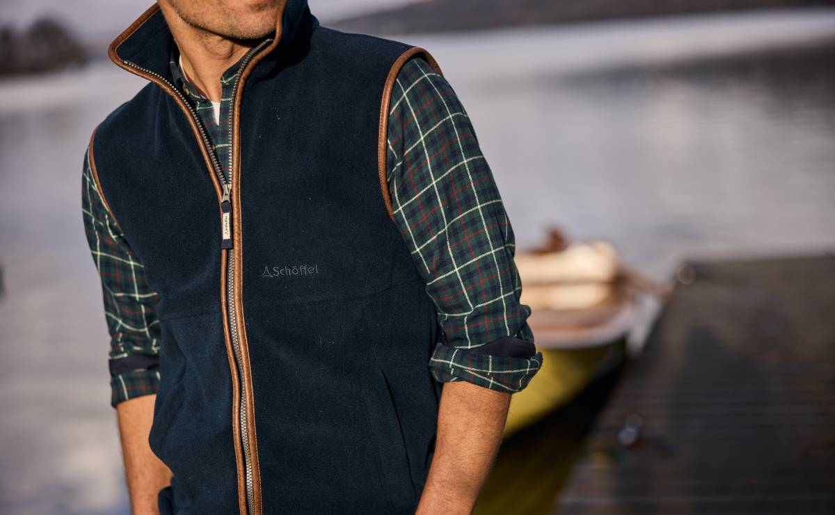 Person wearing a navy gilet over a checked shirt by a body of water.