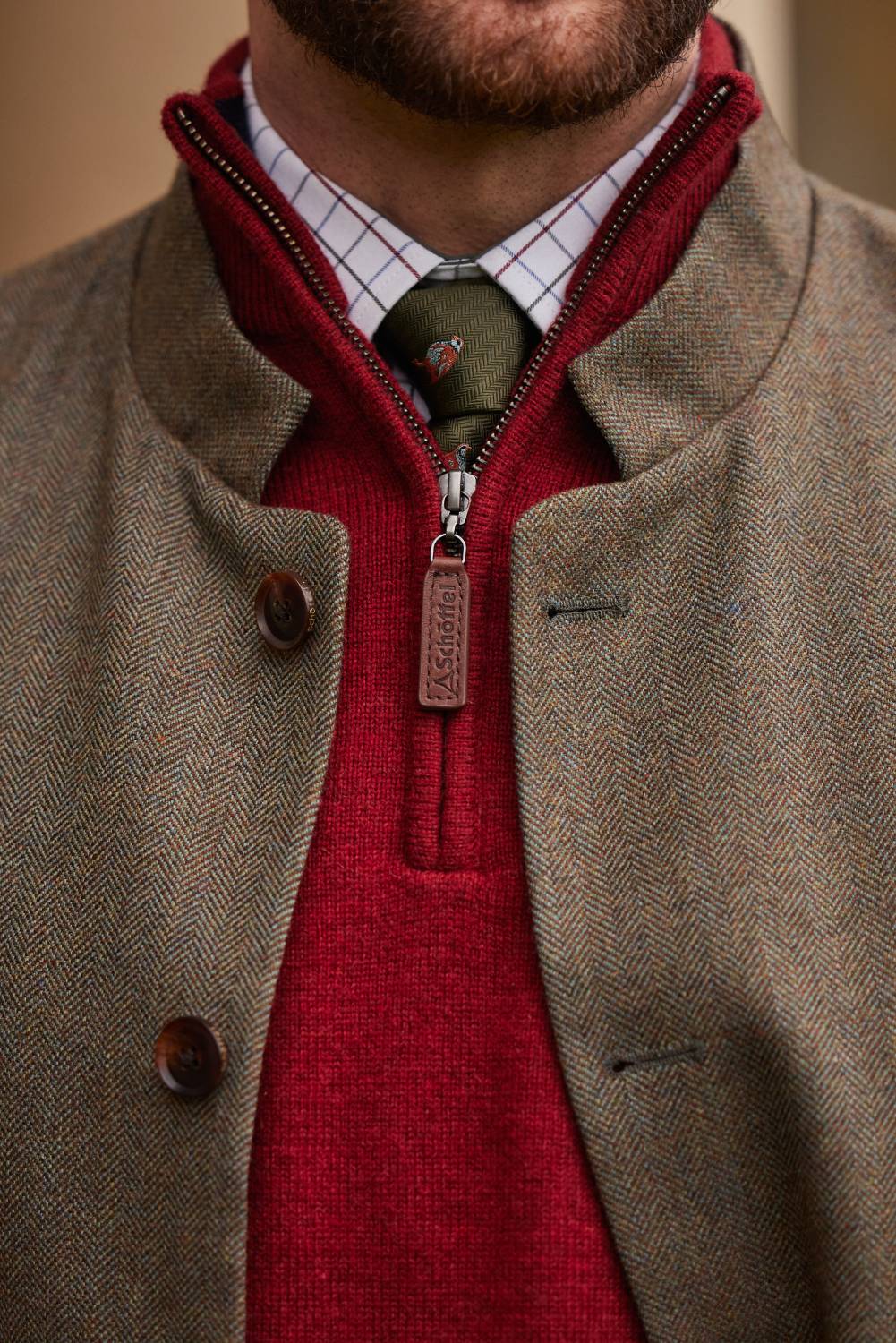 A close-up of a mans collar area. The man wears a tweed waistcoat, red 1/4 zip knitwear, check shirt and a green tie.