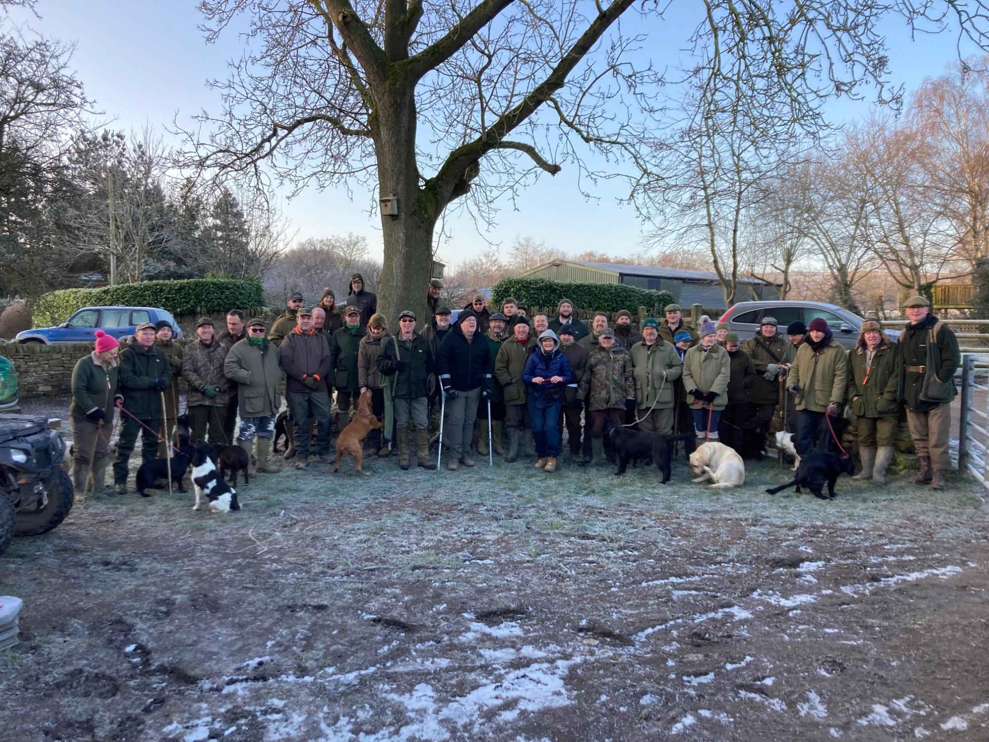 Group of people with dogs standing on a frosty ground with trees and vehicles in the background.