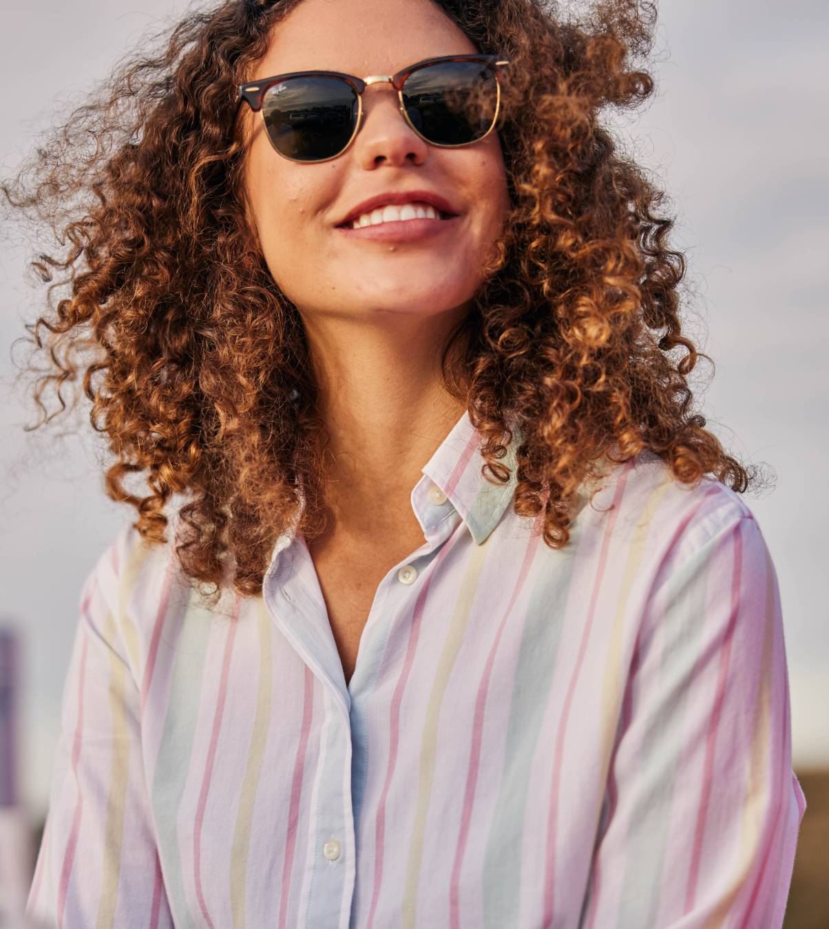 A lady wearing a striped shirt and sunglasses. She is smiling looking away from the camera.