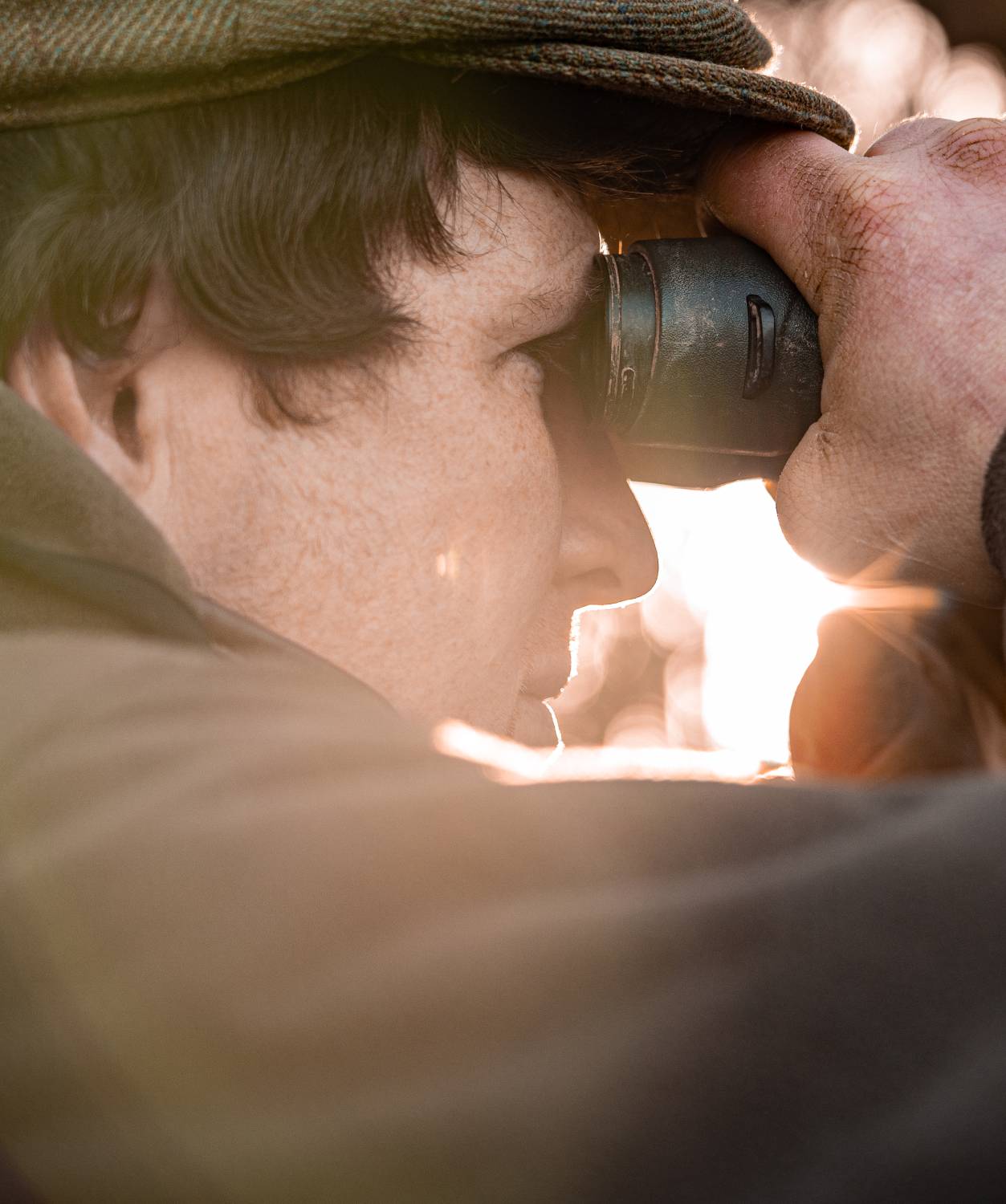 A close-up of a man wearing a brown tweed cap and outdoor jacket, looking through a pair of binoculars. His hands appear weathered, with visible redness and rough skin. The sunlight streams through the background, casting a warm glow on the scene, highlighting the textures of his clothing and the binoculars.