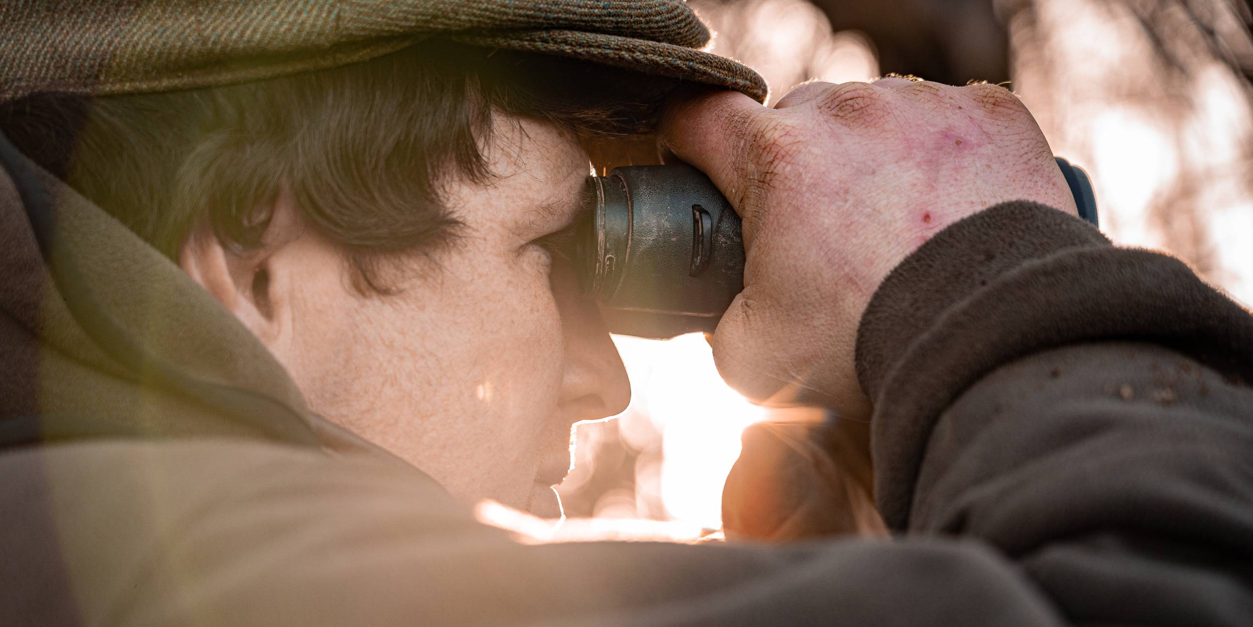 A close-up of a man wearing a brown tweed cap and outdoor jacket, looking through a pair of binoculars. His hands appear weathered, with visible redness and rough skin. The sunlight streams through the background, casting a warm glow on the scene, highlighting the textures of his clothing and the binoculars.