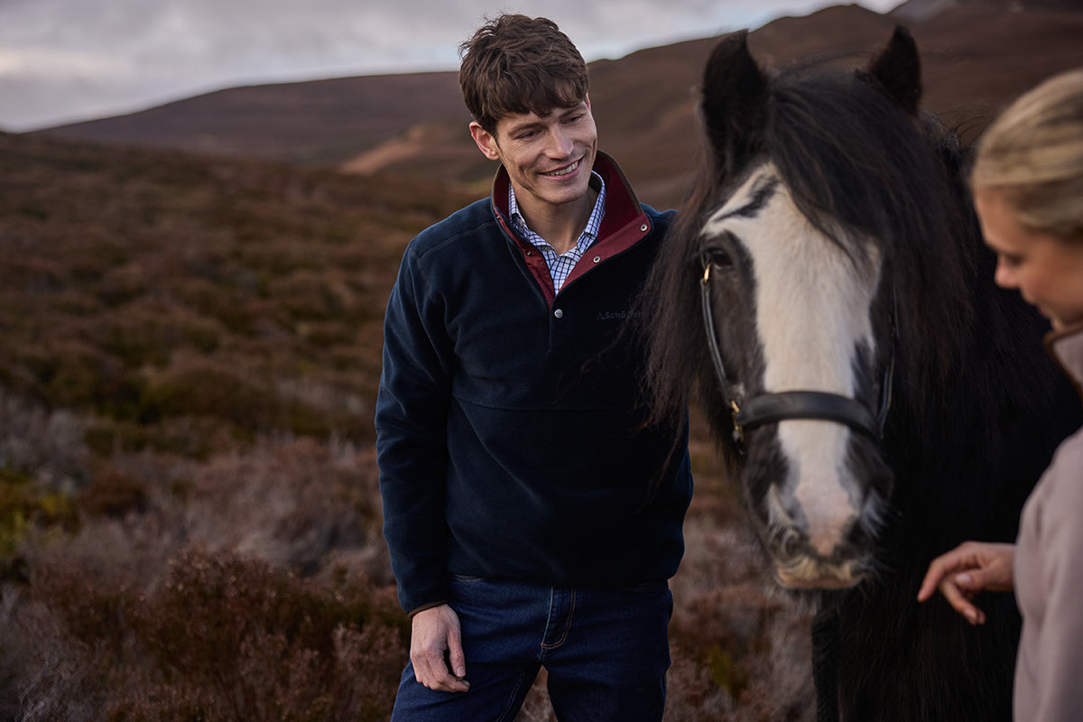 A man in a navy fleece pullover stands outdoors next to a black and white horse.