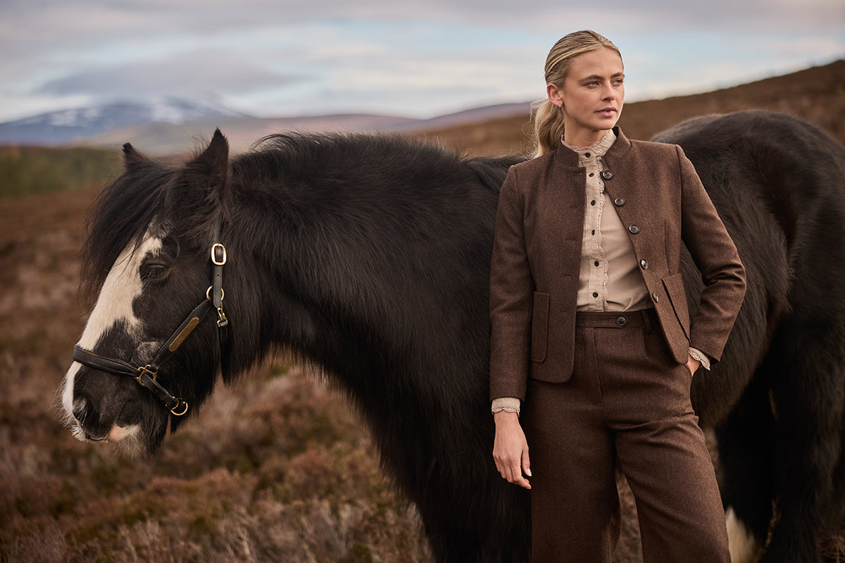 A woman stands outdoors beside a large black horse in a moorland setting,