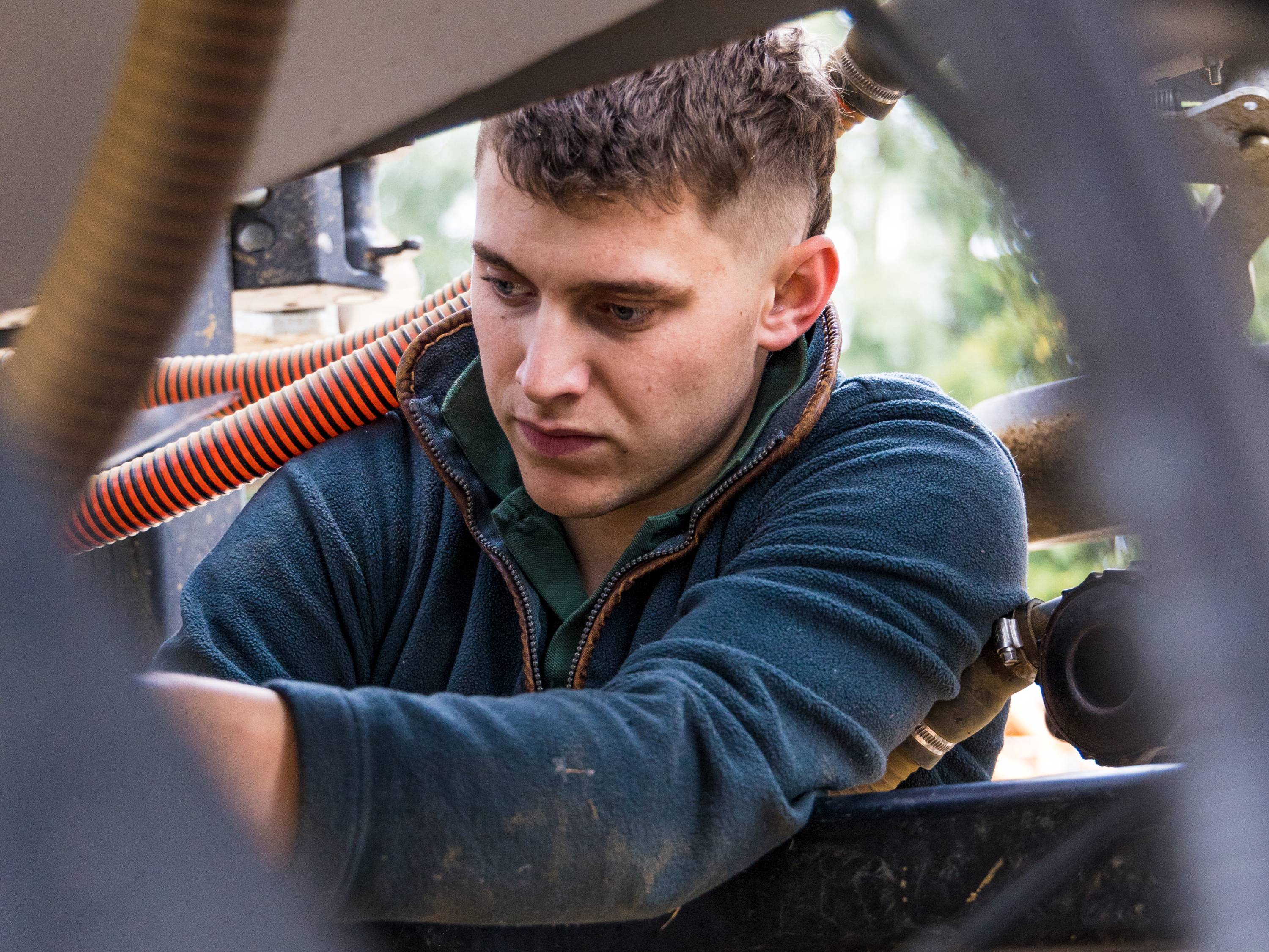 An image of a man wearing a navy jumper and gilet, working on the mechanics of a tractor.