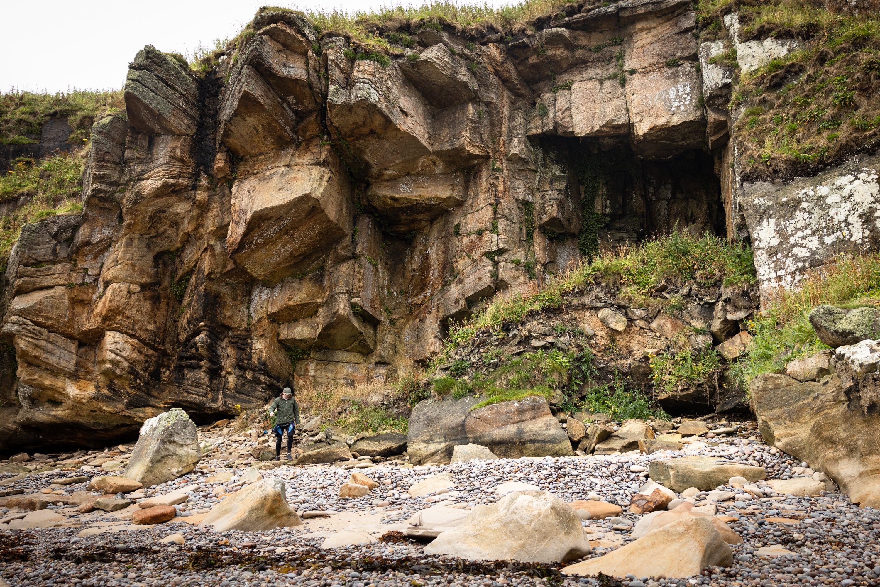 Rocky coastal cliff with grassy patches, pebble beach and person at its base.