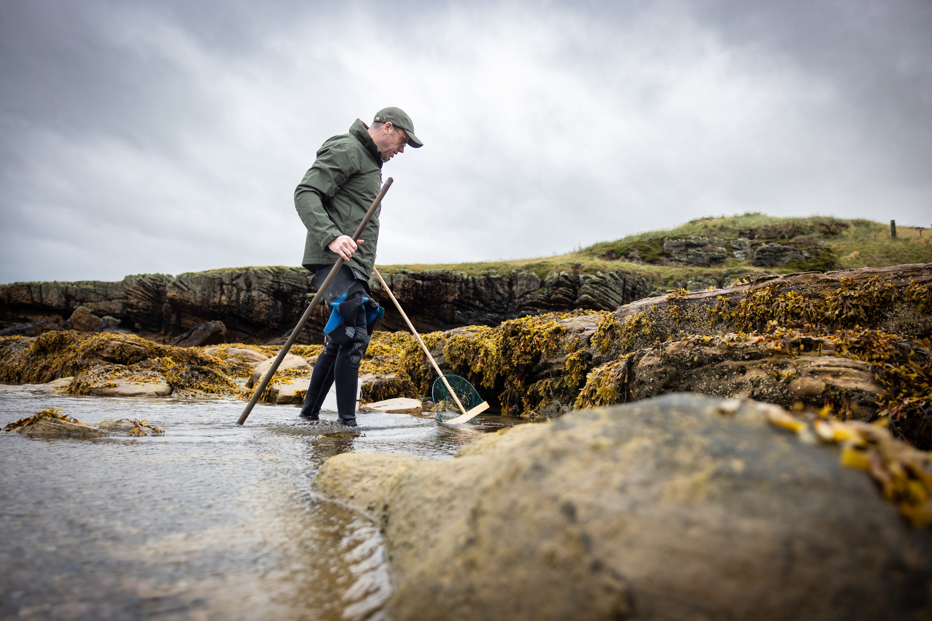 Man in a green jacket and cap stands in shallow water, holding long-handled tools.