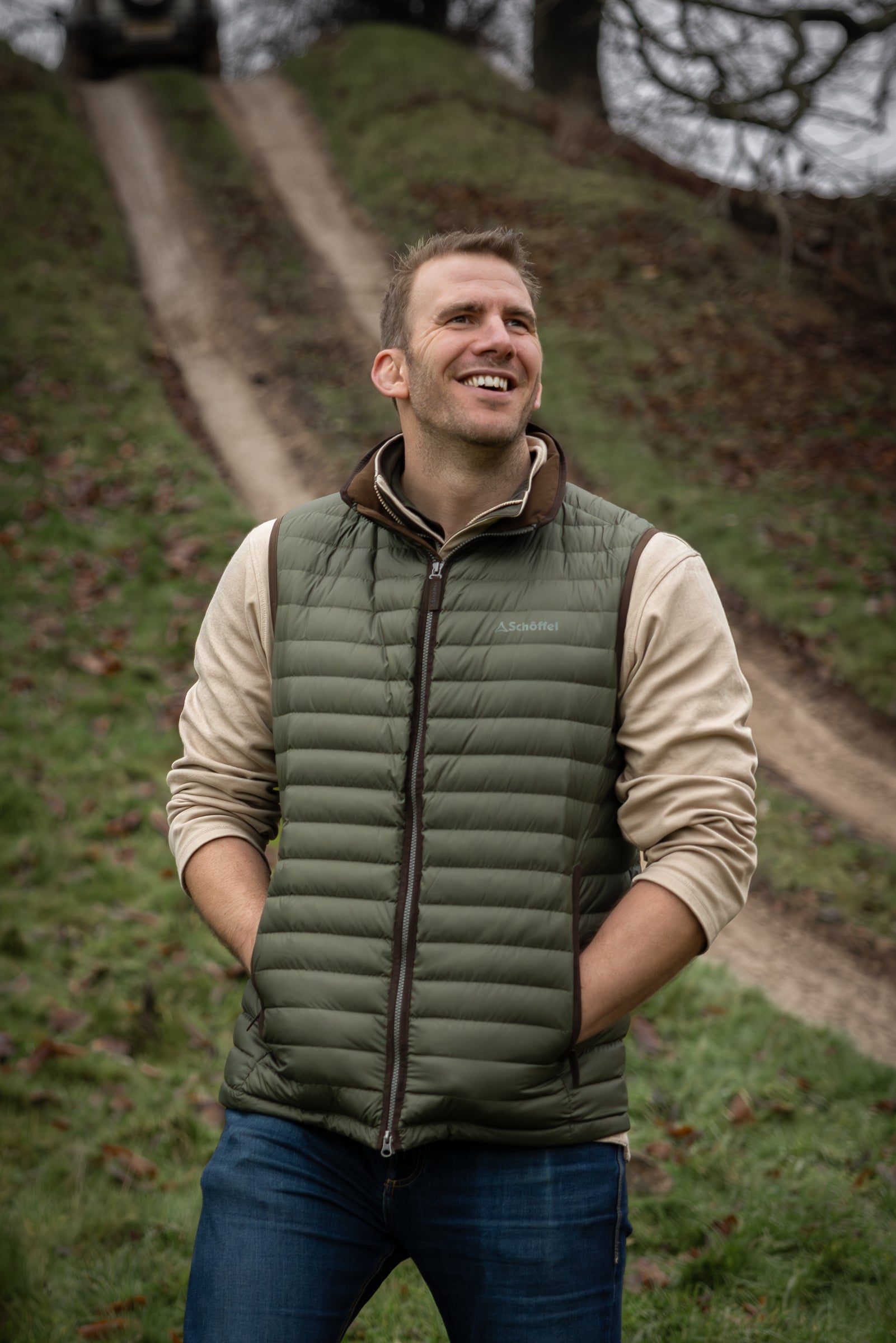 Man wearing a green puffer vest standing outdoors on a path with grass and trees in the background.