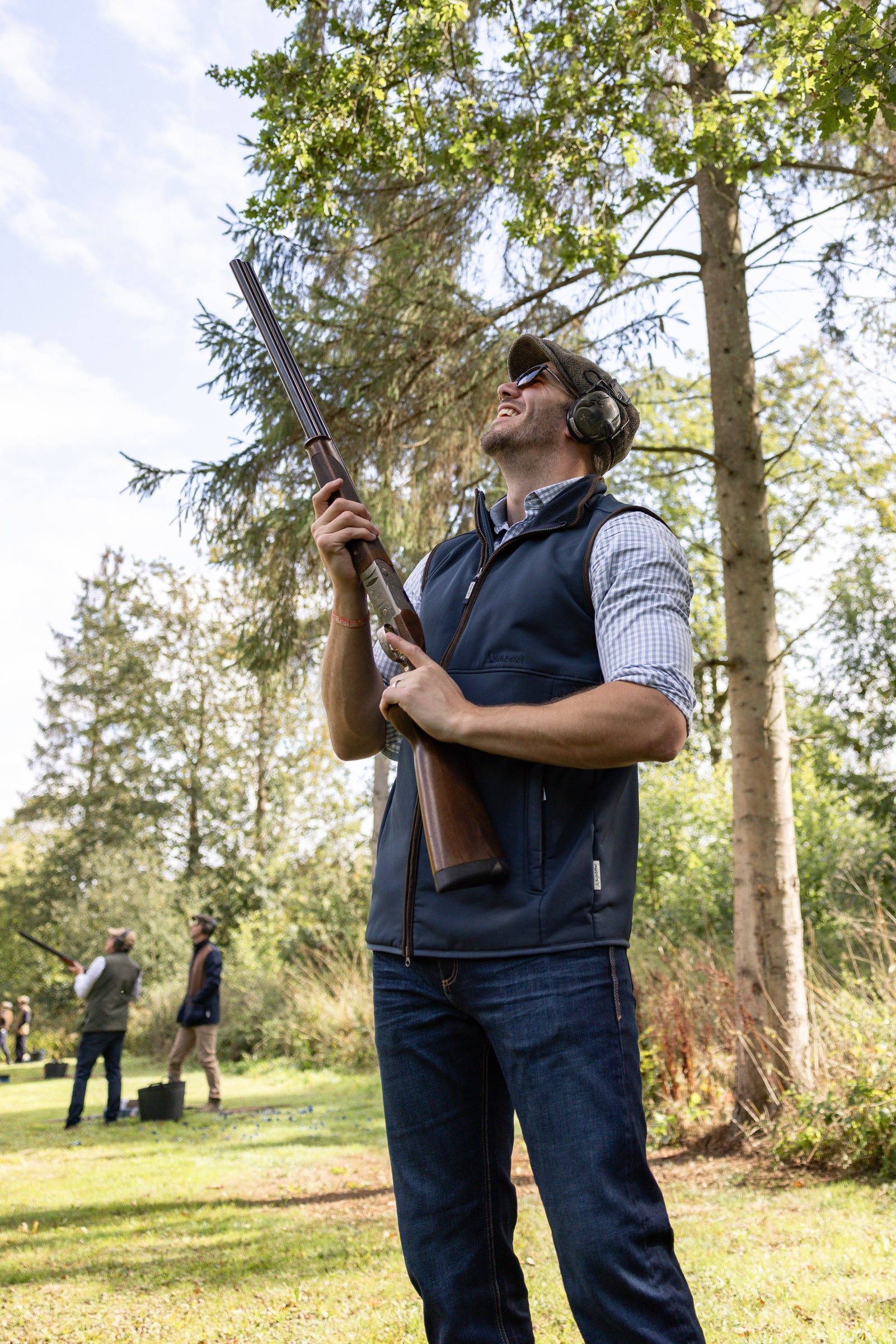 Man holding a rifle in an outdoor setting with trees and people in the background