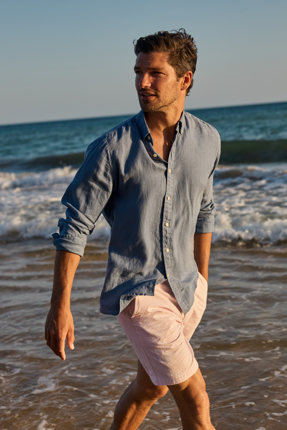 Man walking on a beach wearing a light blue shirt and pink shorts.