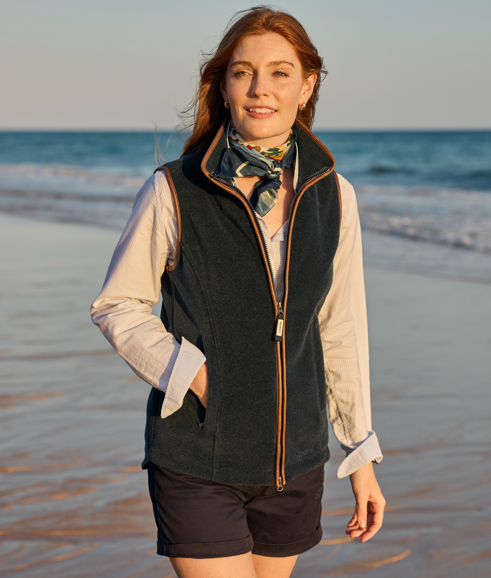 Woman standing on a beach wearing a green vest and black shorts with a scarf around her neck.