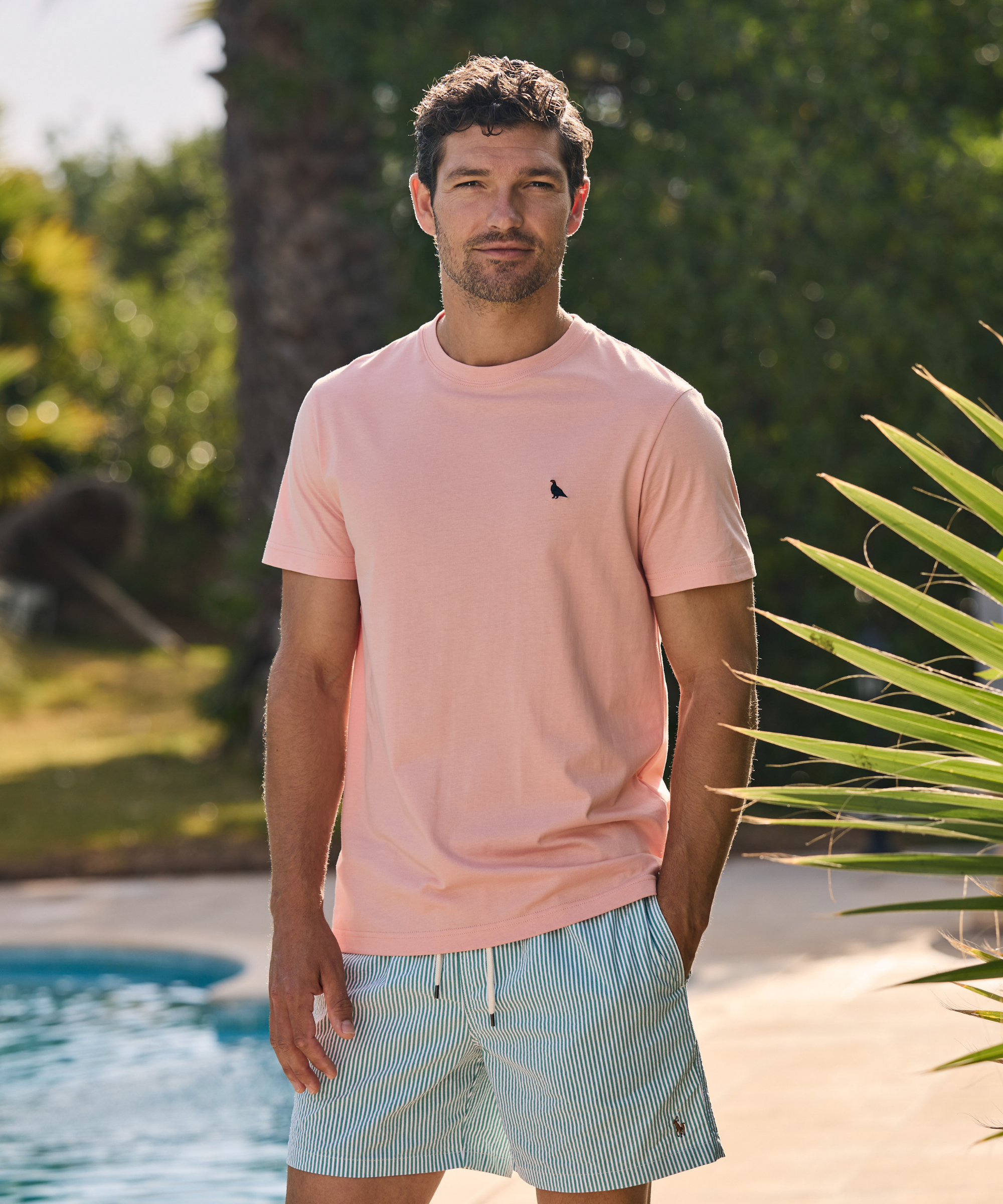 Man wearing a pink t-shirt and light blue shorts standing by a pool with greenery in the background