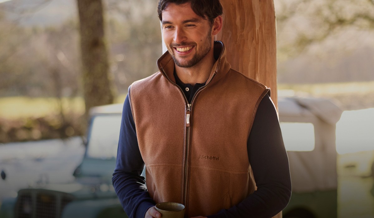 Man wearing a brown leather gilet holding a mug outdoors with a blurred background