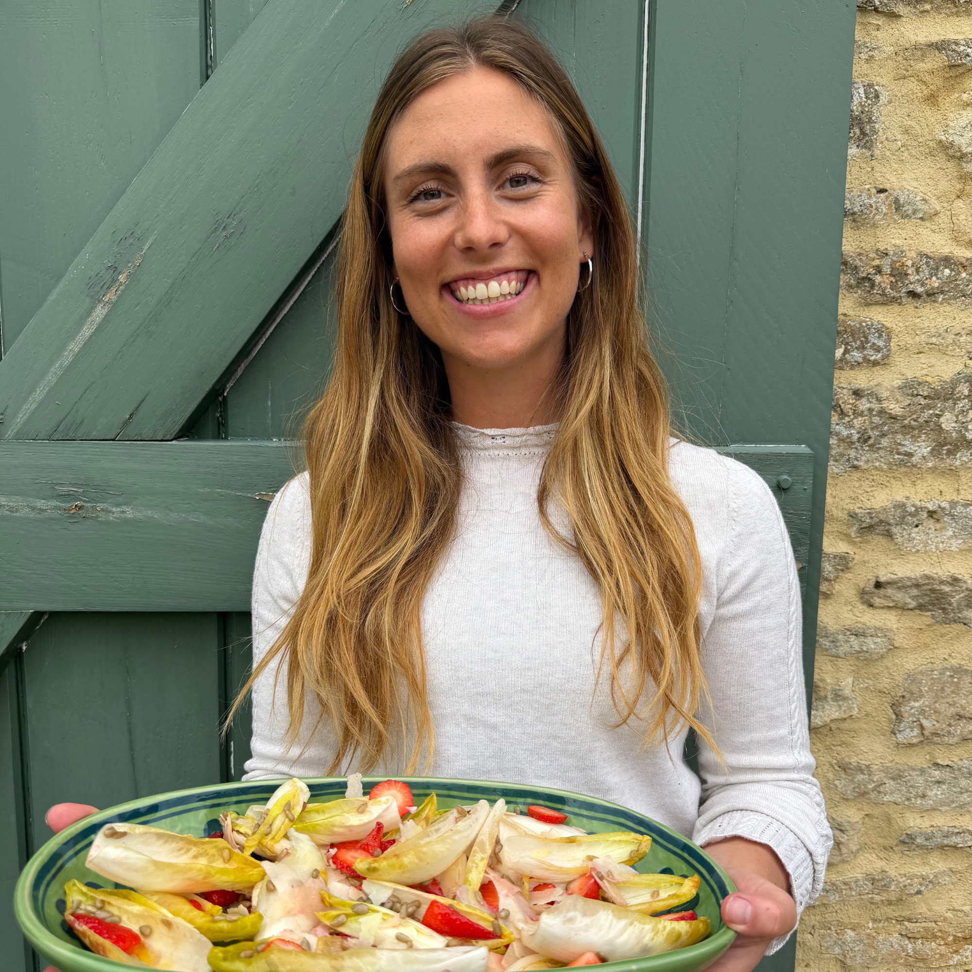 Woman holding a plate of food in front of a green door and stone wall.