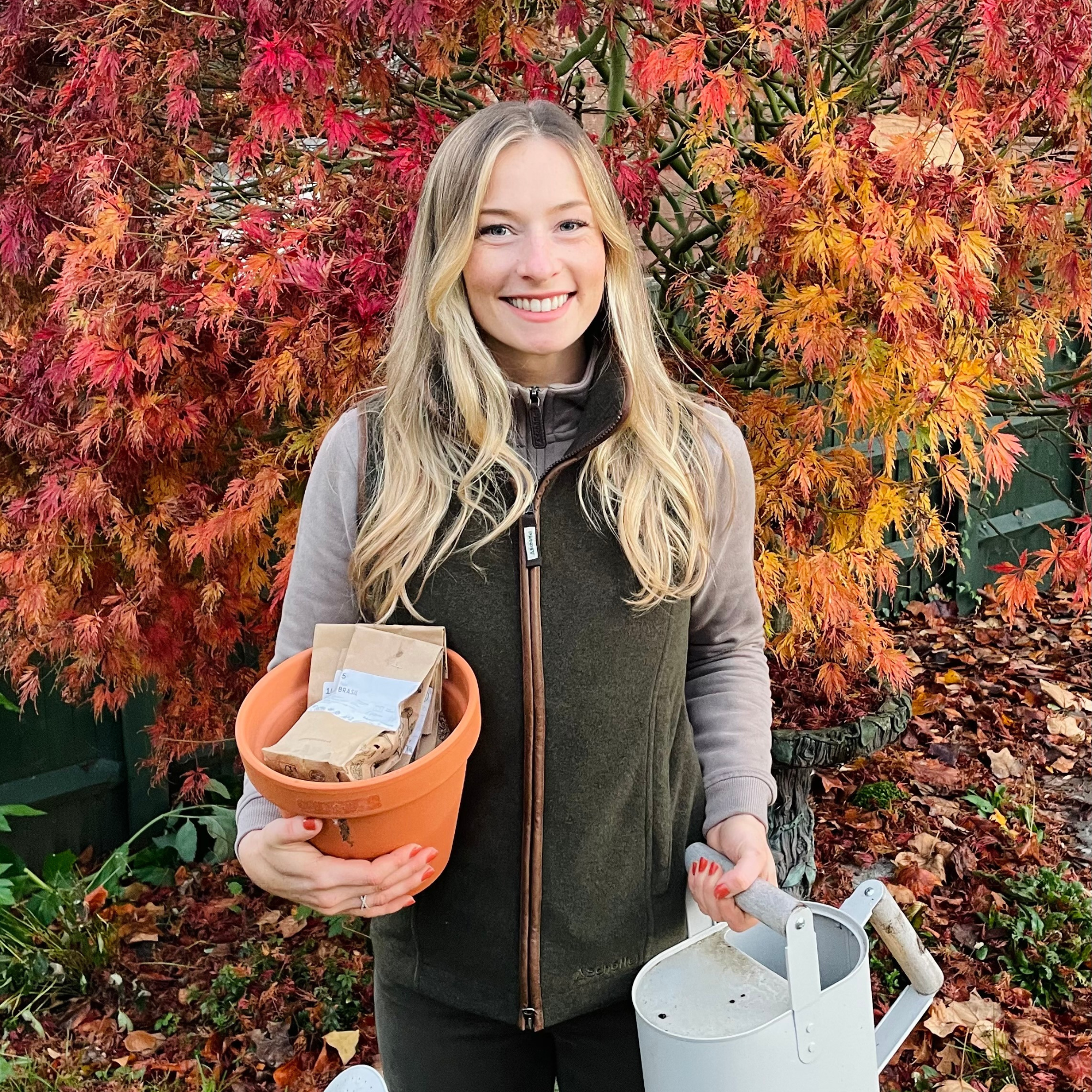 Woman holding a pot with soil and a watering can in front of autumn foliage