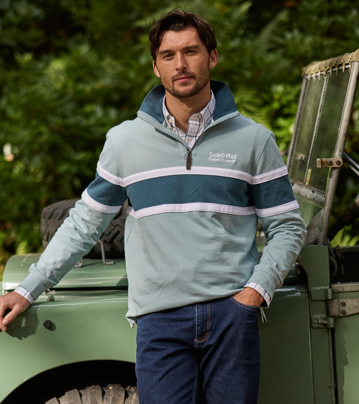 A man wearing a light blue and teal quarter-zip pullover jumper with white stripes and the Schöffel logo, standing outdoors next to a green vehicle.