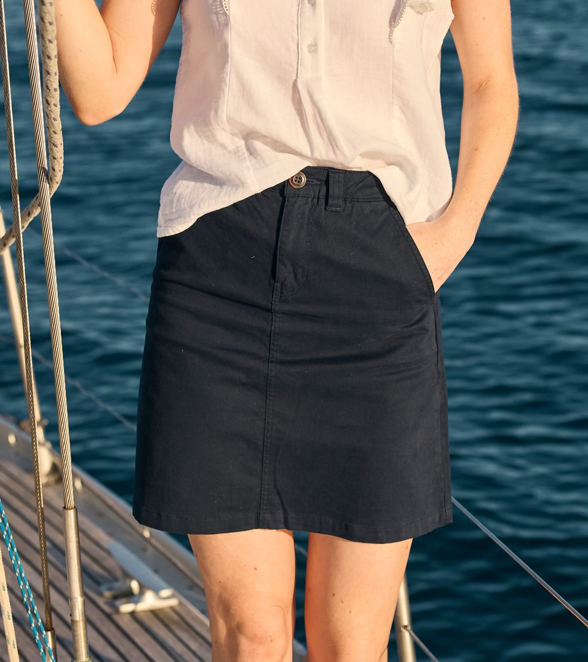 A woman wearing a navy blue mini skirt and a white sleeveless top, standing on a boat with blue water in the background.