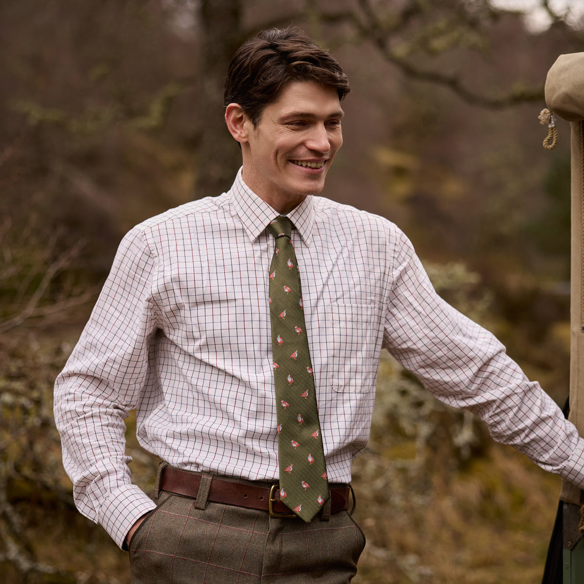 A man wearing a light pink and white checkered long-sleeve shirt, a green patterned tie, and brown tweed trousers, standing outdoors against a blurred natural background.