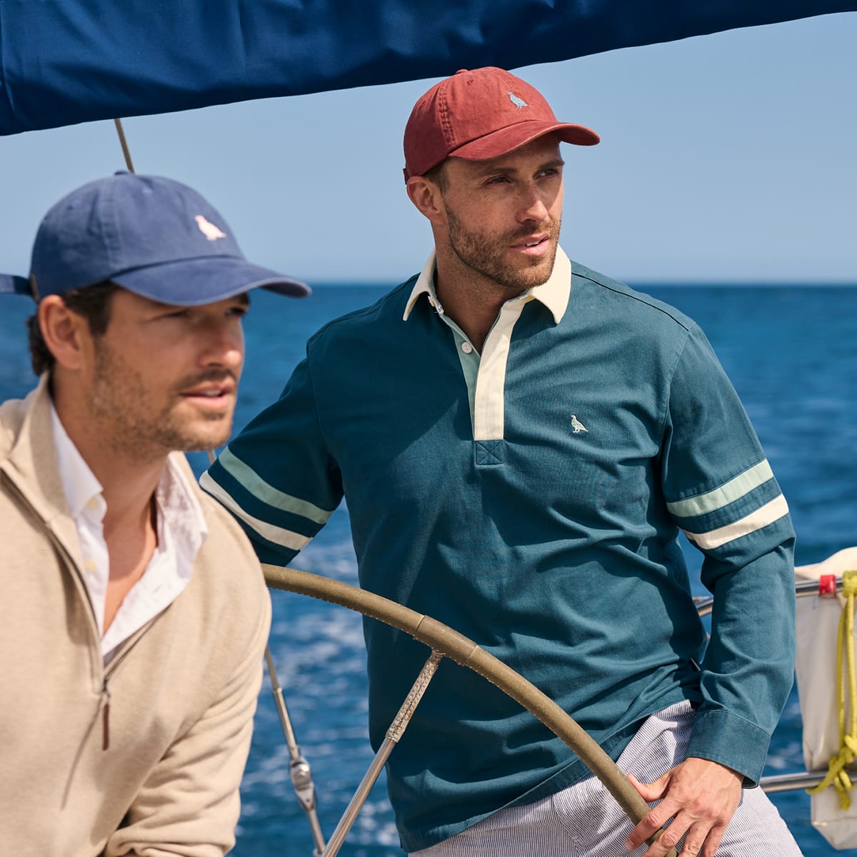 Two men sailing on a boat with the ocean in the background, wearing Nautica branded apparel. One man wears a teal long-sleeve deck shirt with cream trim and a red cap, while the other wears a beige zip-up sweater and a blue cap.