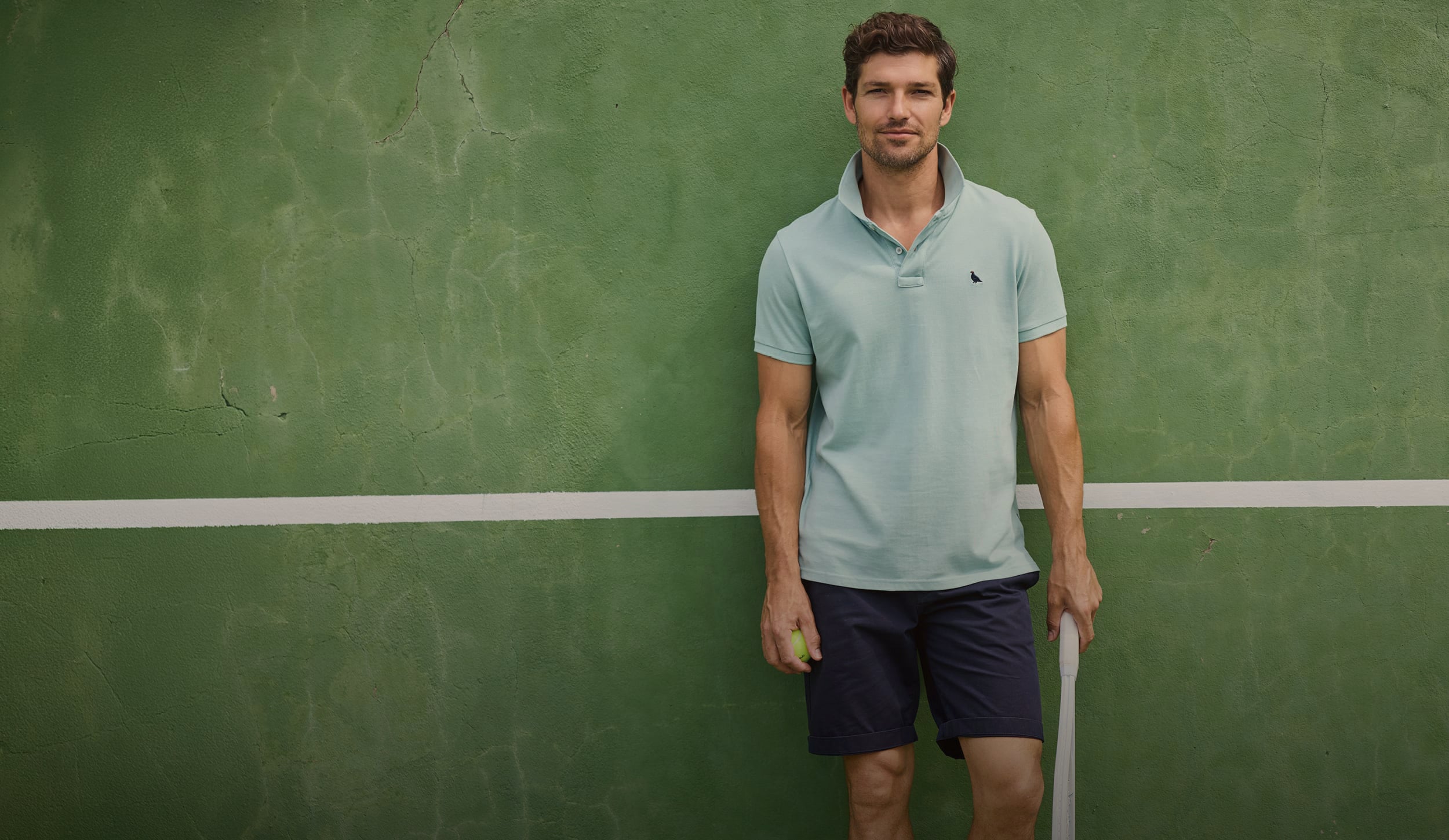 A man stands on a green tennis court wearing a light blue polo shirt and navy shorts, holding a tennis racket and a tennis ball.