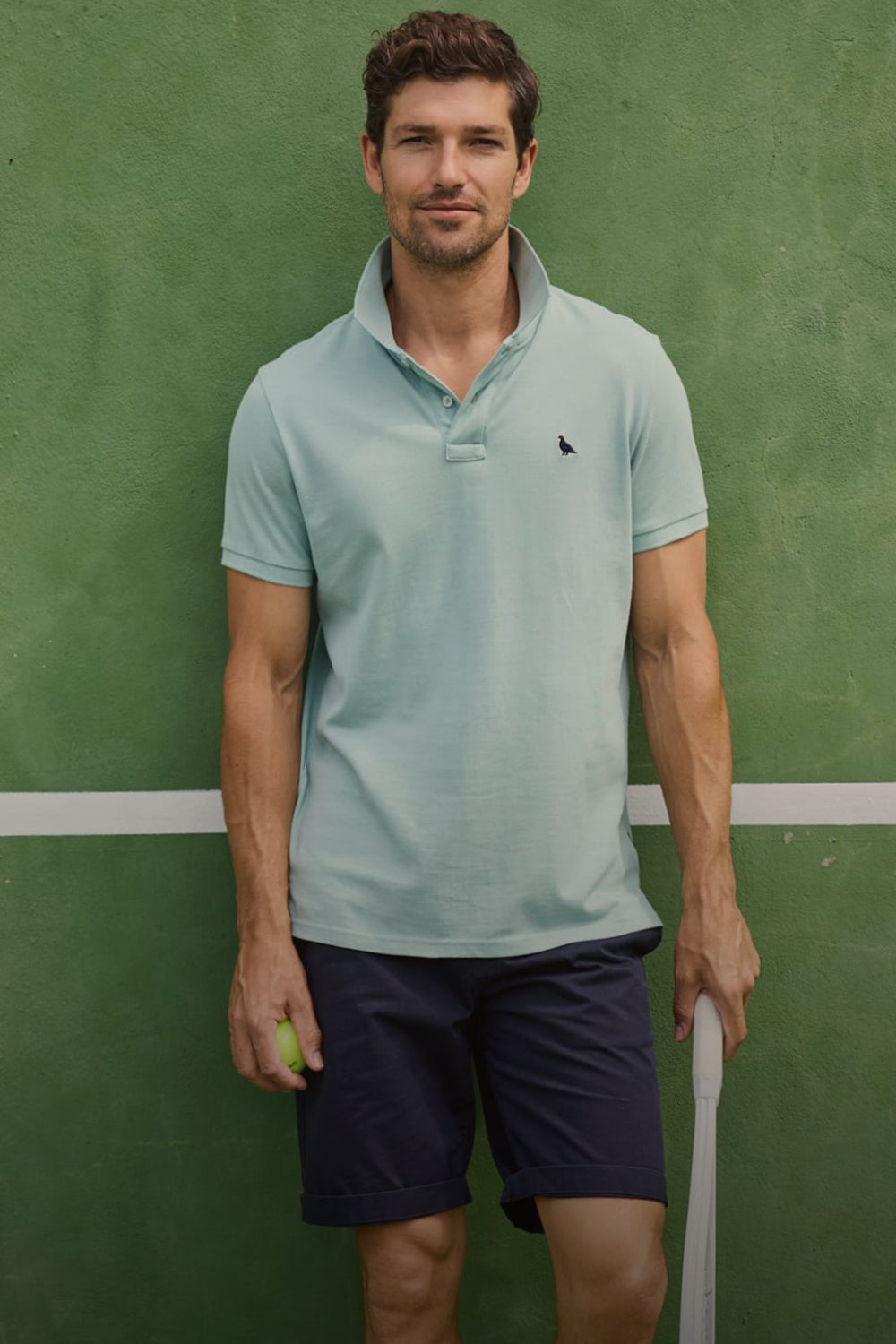 A man wearing a light sage green polo shirt and navy shorts stands against a green tennis court wall holding a tennis racket and a tennis ball.