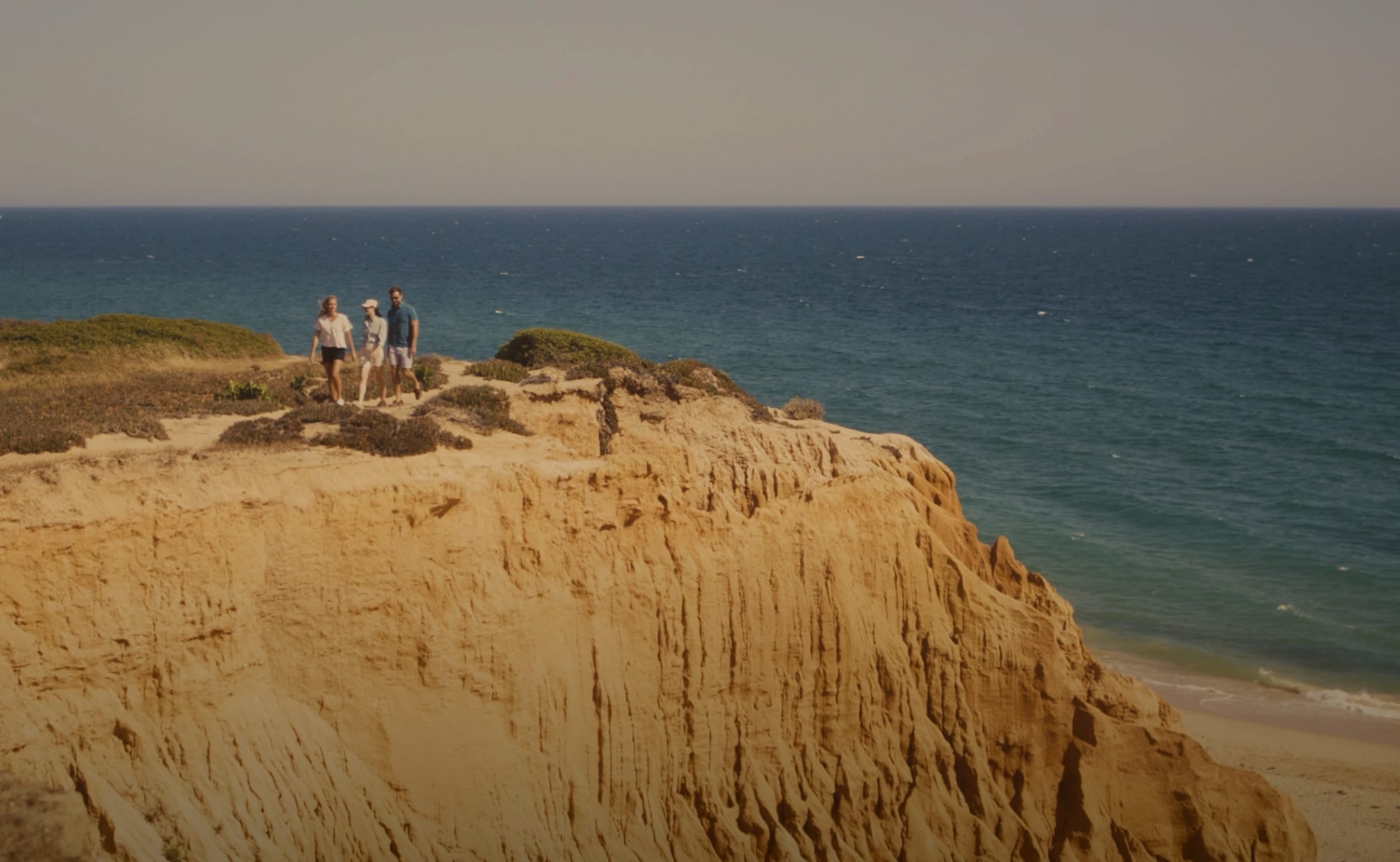 A scenic coastal landscape with tall sandy cliffs overlooking a blue ocean, with three small people standing on the cliff edge.