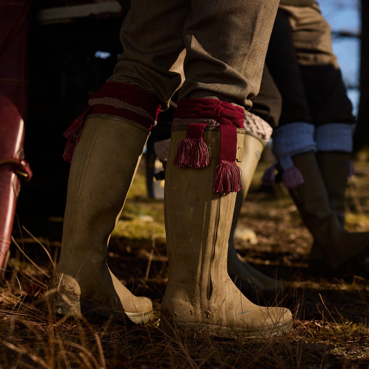 Close-up of a person wearing tall wellington boots with red shooting socks featuring tassels and matching red cuffs, standing outdoors on grassy ground with other people and boots blurred in the background.
