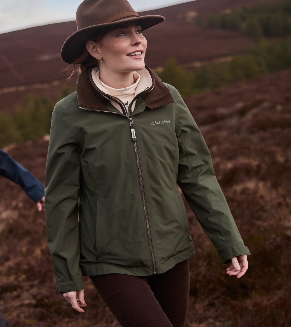 A woman wearing an olive green outdoor jacket with a brown collar, a brown wide-brim hat, and brown pants, walking in a moorland landscape.