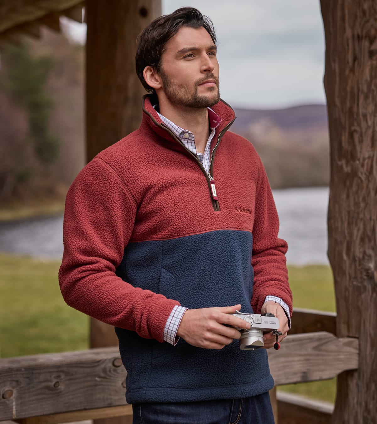 A man wearing a two-tone fleece in red and navy blue, standing outdoors near a wooden structure with a lake and hills in the background.