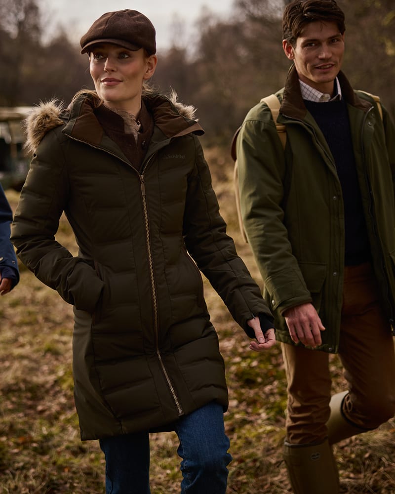A woman wearing a dark olive green mid-length puffer parka with a fur-trimmed hood and a brown flat cap, walking outdoors next to a man in an olive green jacket.