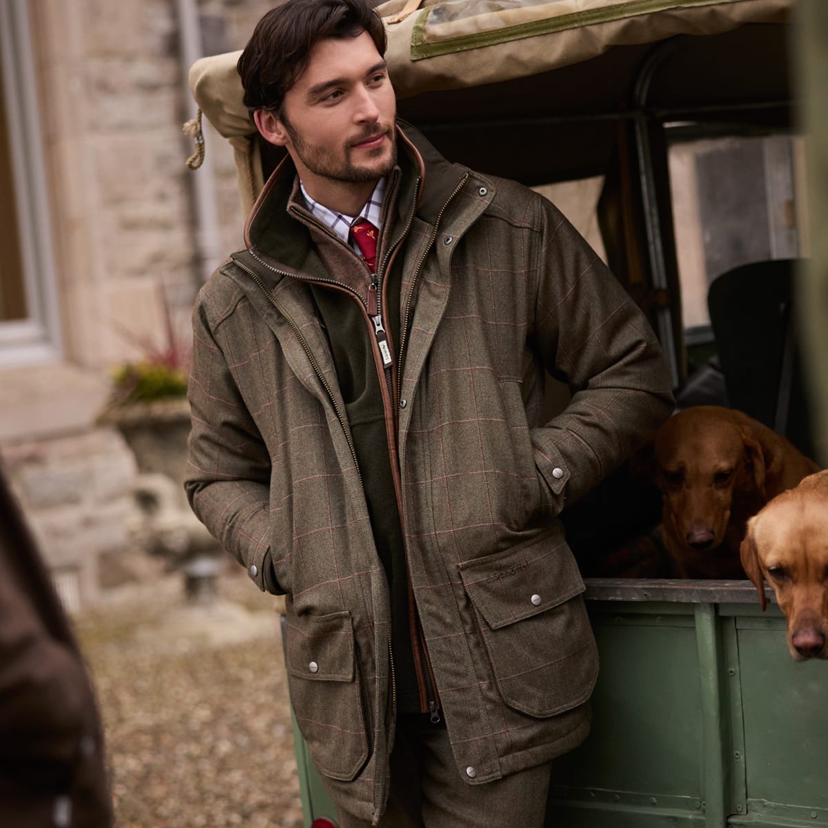 Man wearing green tweed jacket stands next to a green vehicle with two dogs