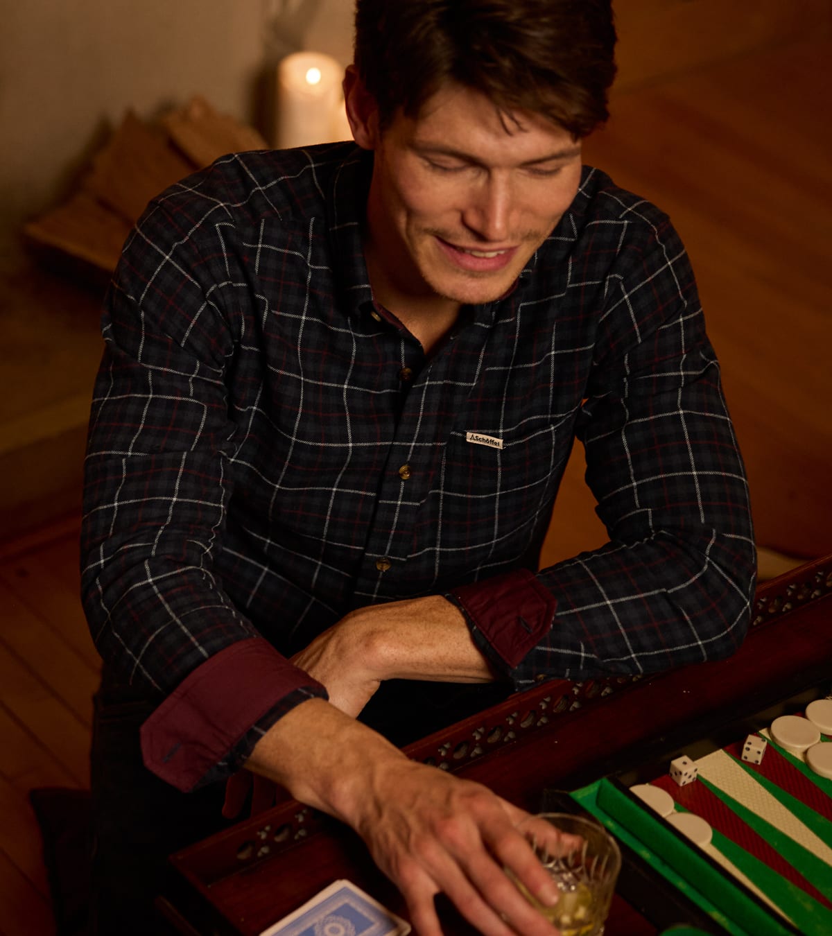 Man in navy check flannel shirt with burgundy cuff lining playing backgammon by candlelight