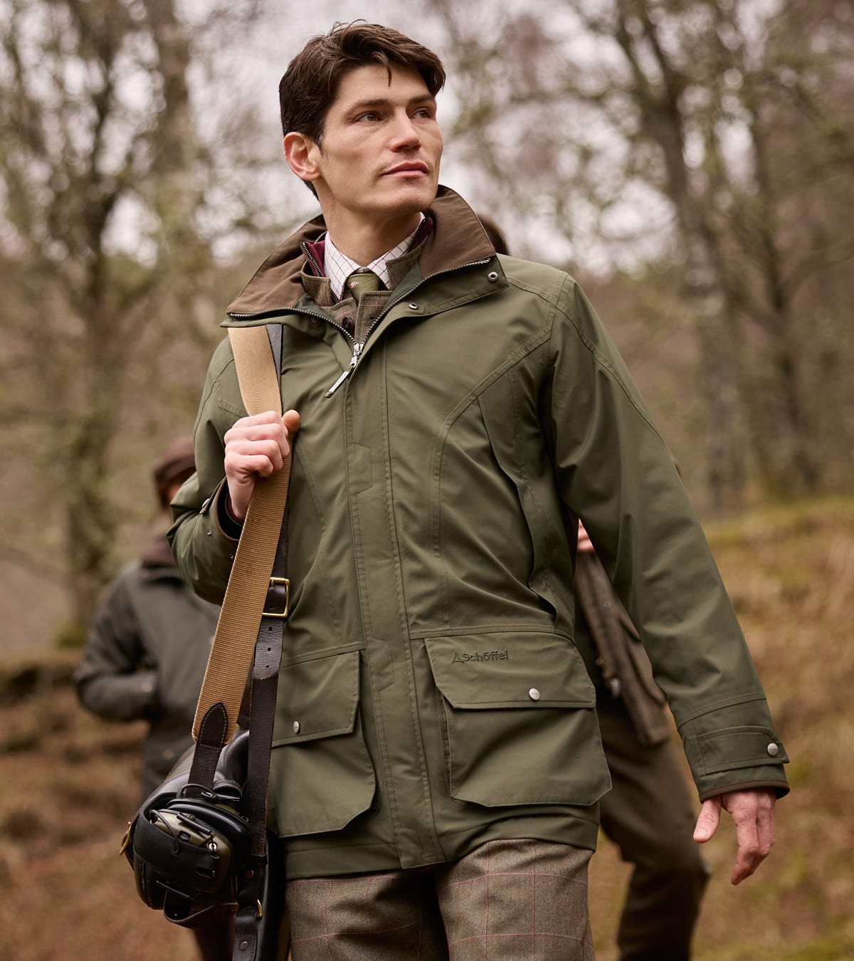 Man in olive green shooting jacket with brown corduroy collar carrying gun slip and ear defenders