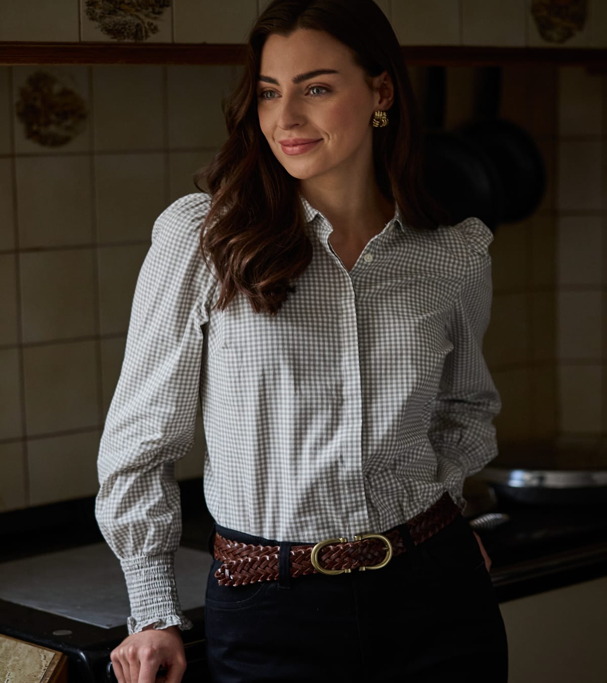 Woman in grey gingham check shirt with ruffled cuffs and woven leather belt in a kitchen setting