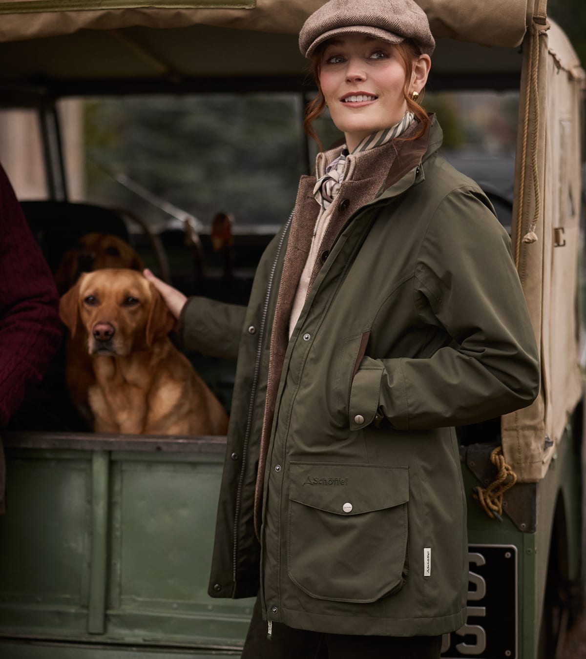 Woman in olive green shooting jacket and tweed flat cap petting a dog beside a vintage Land Rover