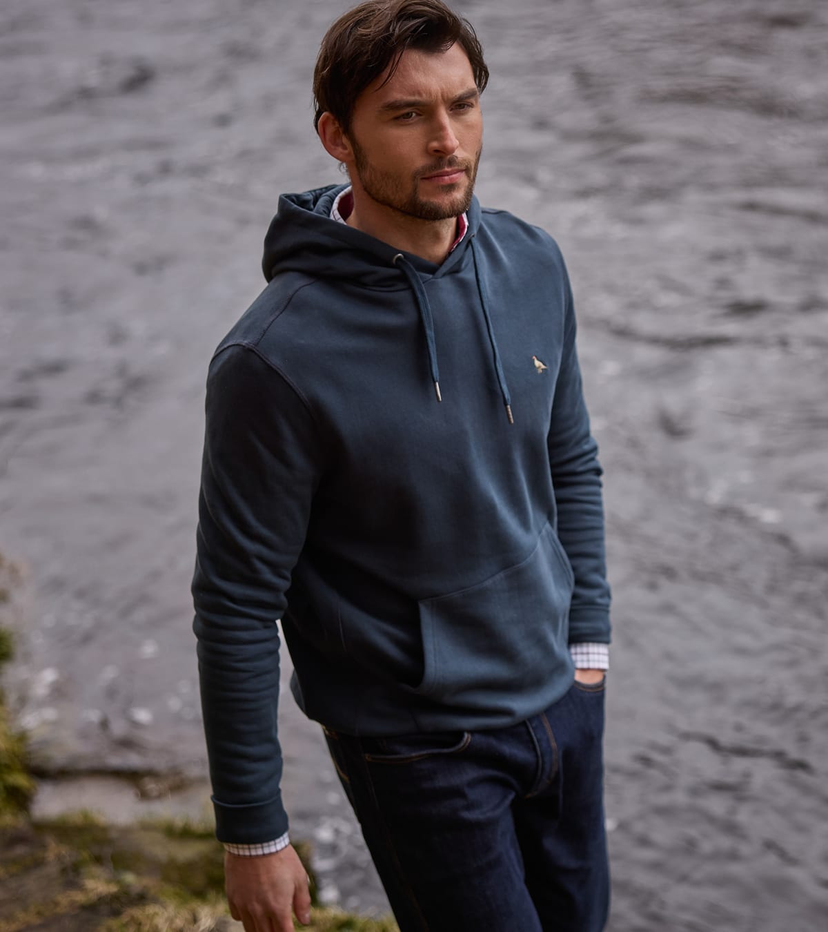 A man wearing a navy hoodie with a small embroidered bird logo standing near a river
